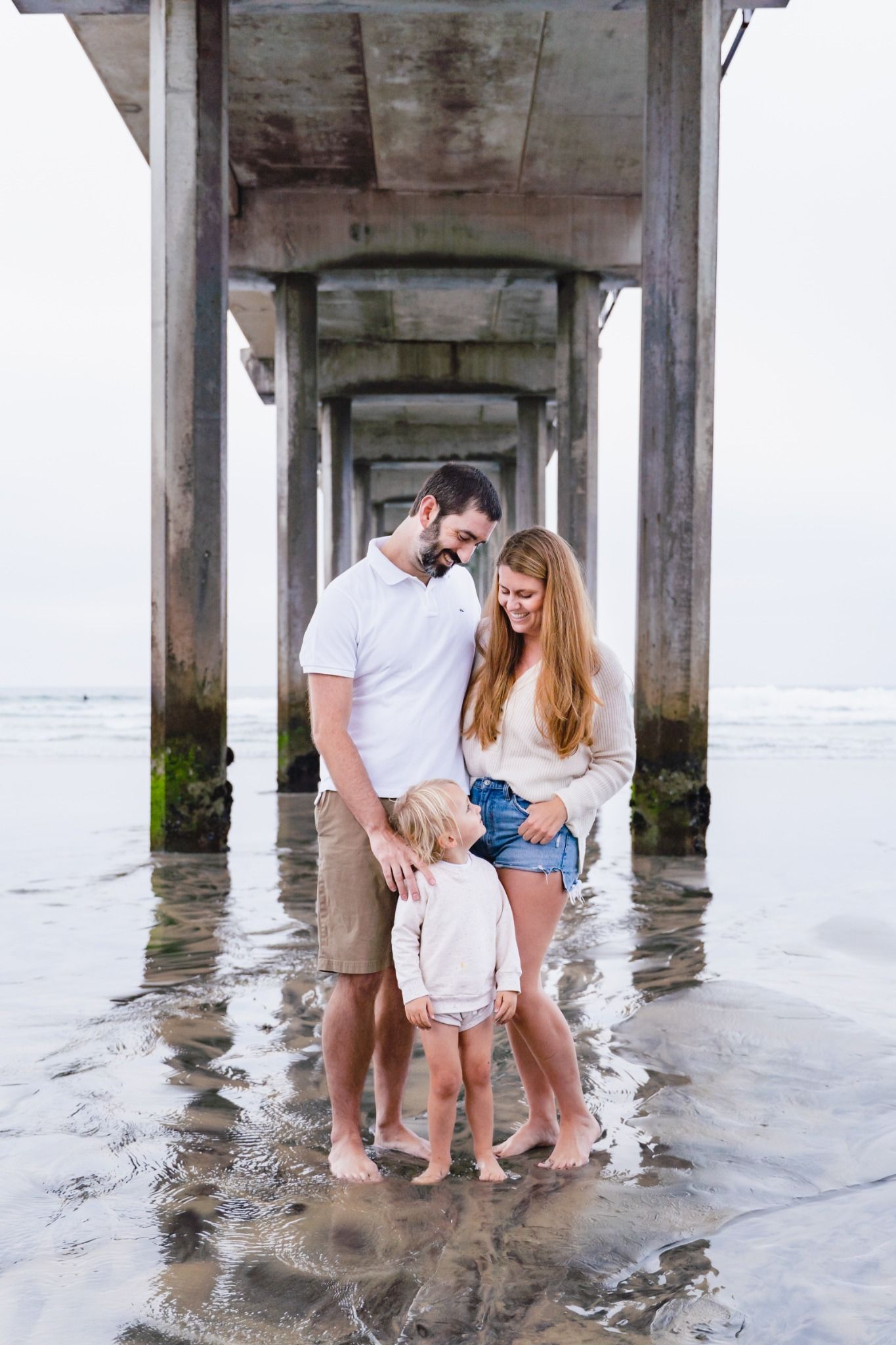 Family of three standing barefoot under a beach pier in shallow water, parents smiling at their young child.