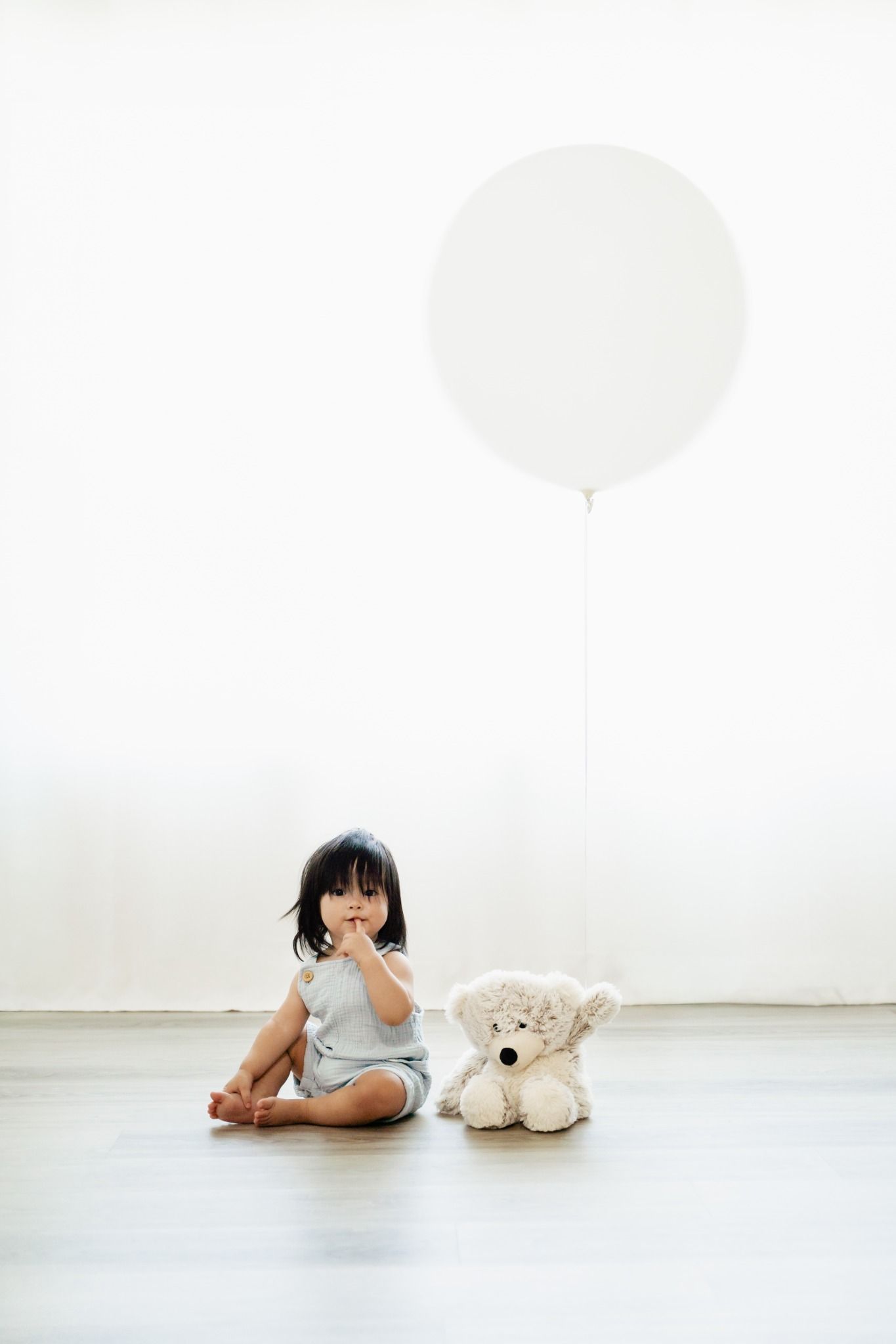 Young Asian toddler girl sitting on floor with white teddy bear and large white balloon against bright white wall.