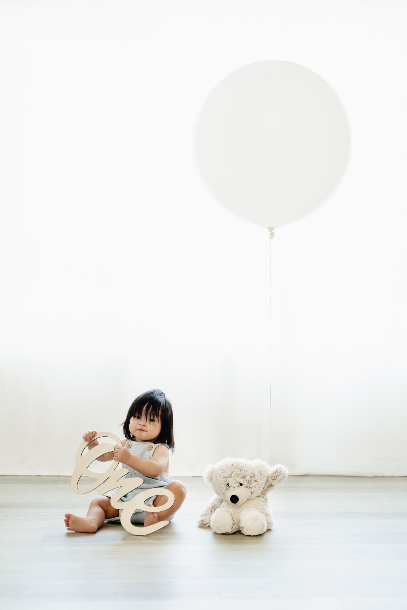 Toddler girl sitting on floor with wooden toy and teddy bear next to large white balloon against white wall.