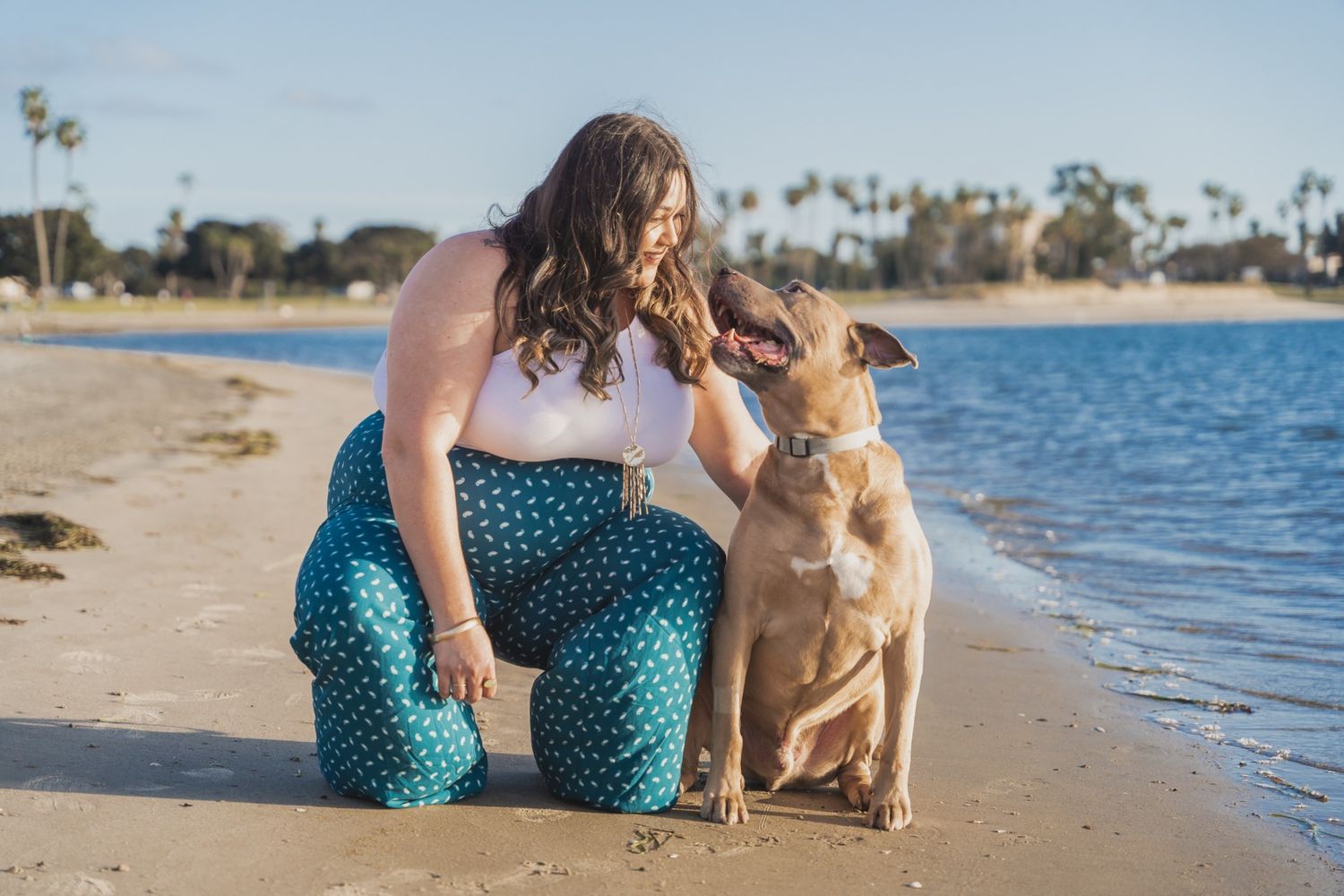 Plus-size woman in teal pants bonding with her tan dog on a sunny beach.