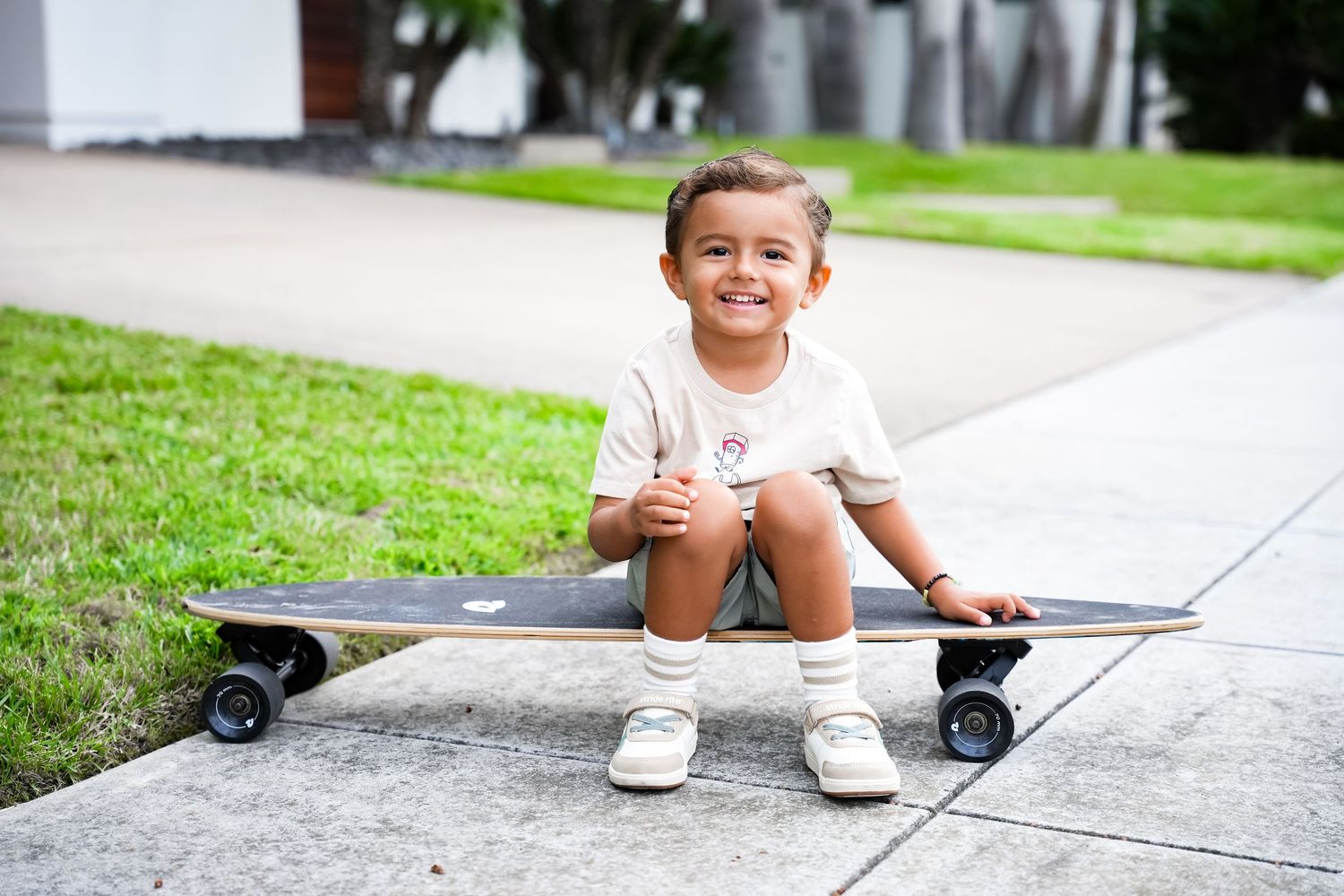 Smiling toddler girl sitting on black skateboard on suburban sidewalk.
