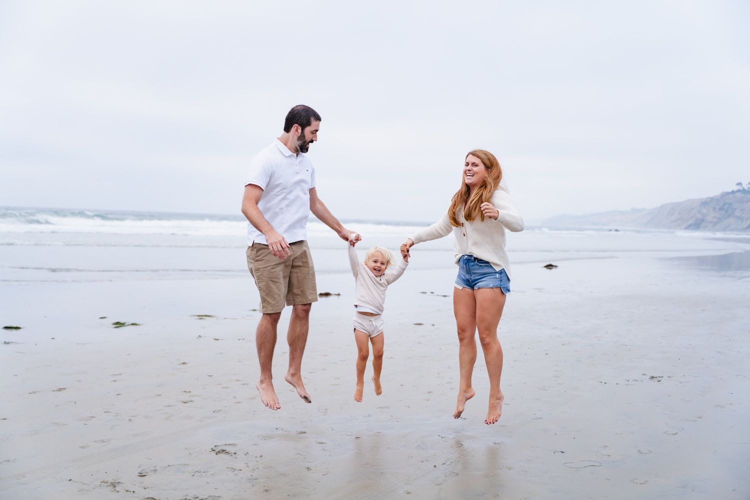 Family of three enjoying a beach day, parents swinging toddler by hands on sandy shore with ocean waves behind.