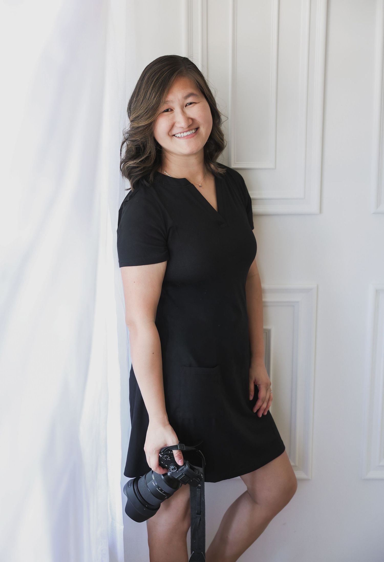 Professional female photographer in black dress smiling while holding camera, posing by white door.