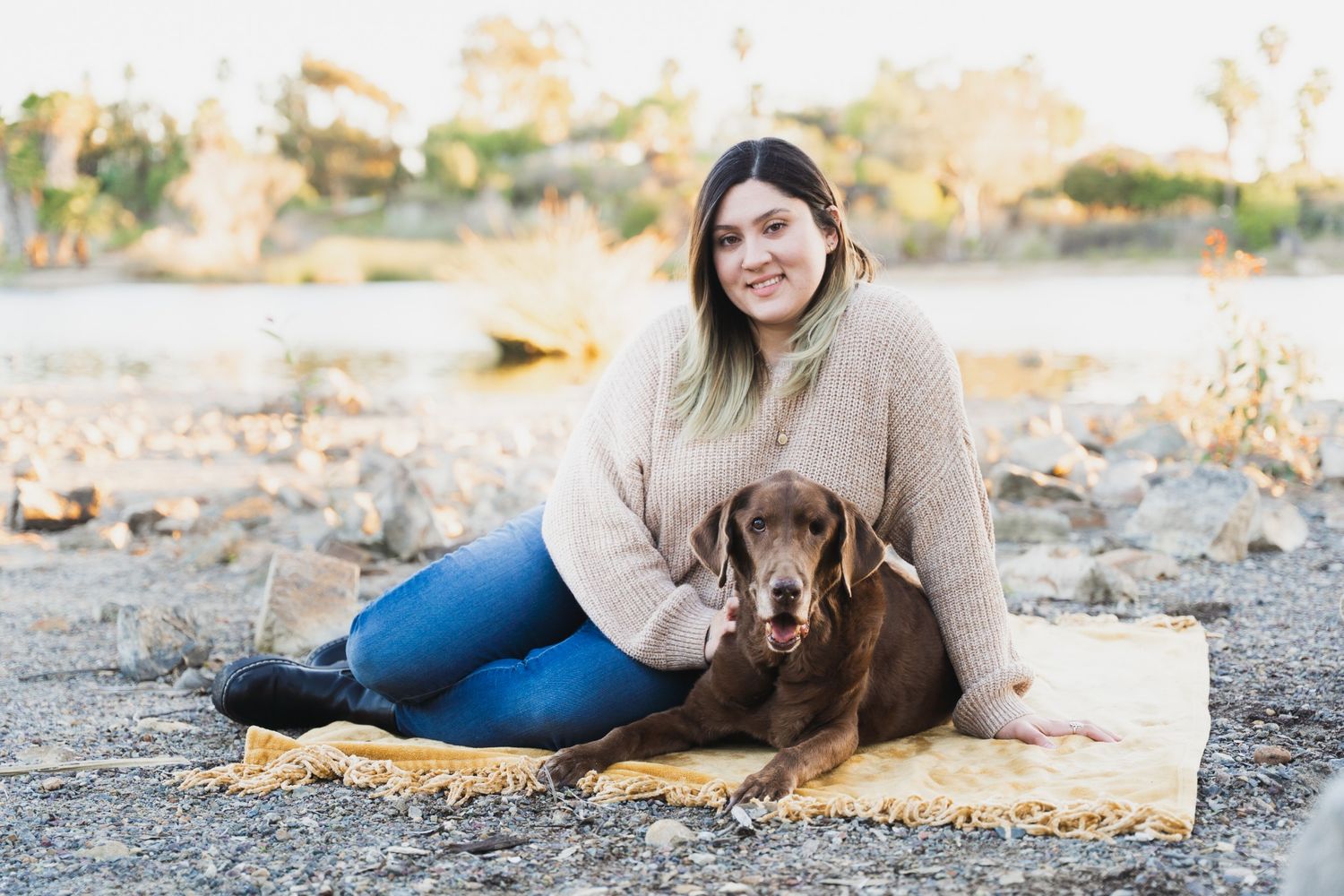Woman sitting on yellow blanket outdoors with chocolate Labrador dog, smiling in cozy sweater and jeans.
