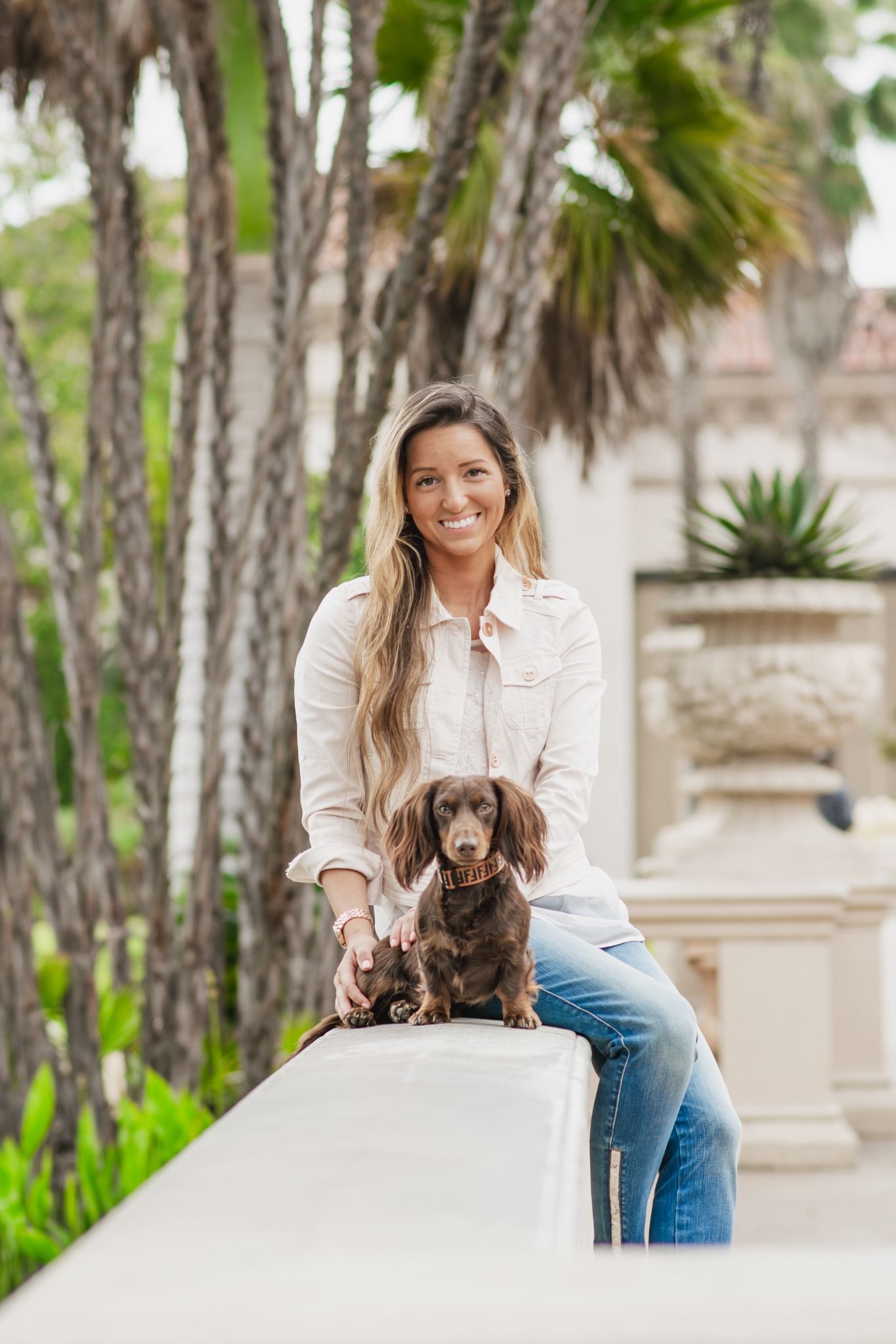 Smiling blonde woman posing with chocolate Dachshund dog on white ledge surrounded by tropical palm trees.