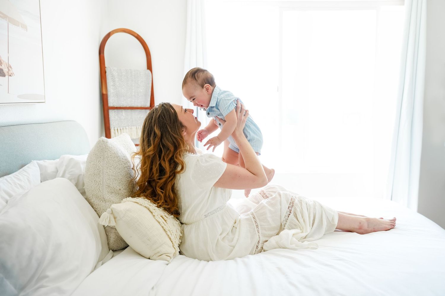 A mother lies on a white bed, joyfully lifting her smiling baby in a bright, airy bedroom.