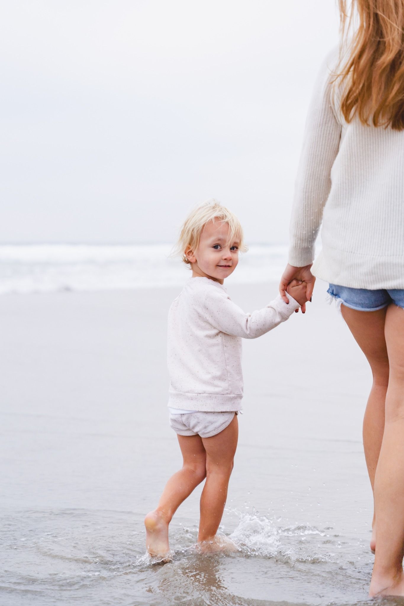 Toddler holding hands with mother at the beach, smiling while standing in shallow ocean waves.
