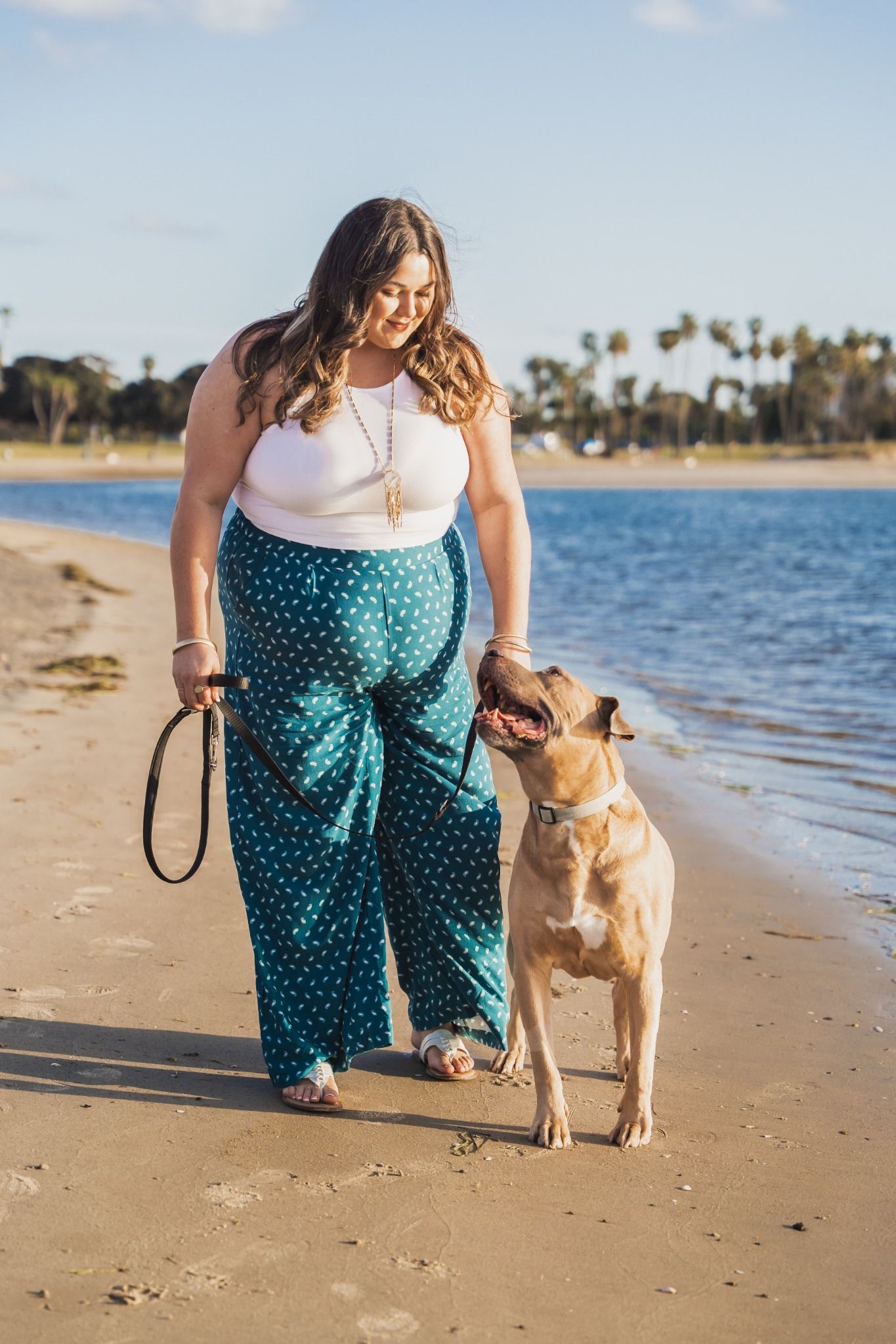 Plus-size woman in teal wide-leg pants walks her tan dog on a sunny beach near calm blue water.