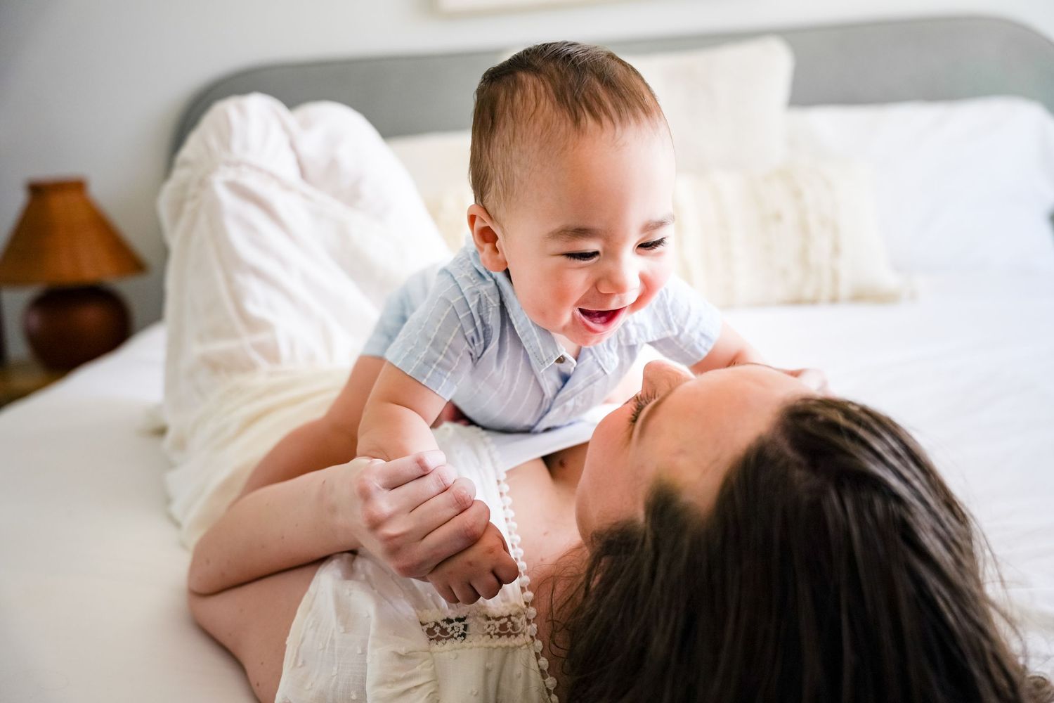 A joyful baby laughs while being held up by a mother lying on a white bed in a bright, cozy room.