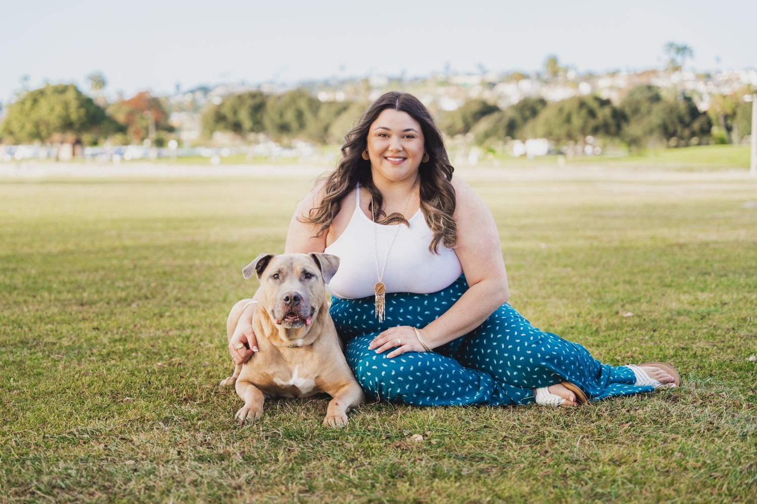 Woman smiling with her tan bulldog on green grass in a sunny park, wearing a white top and blue polka dot pants.