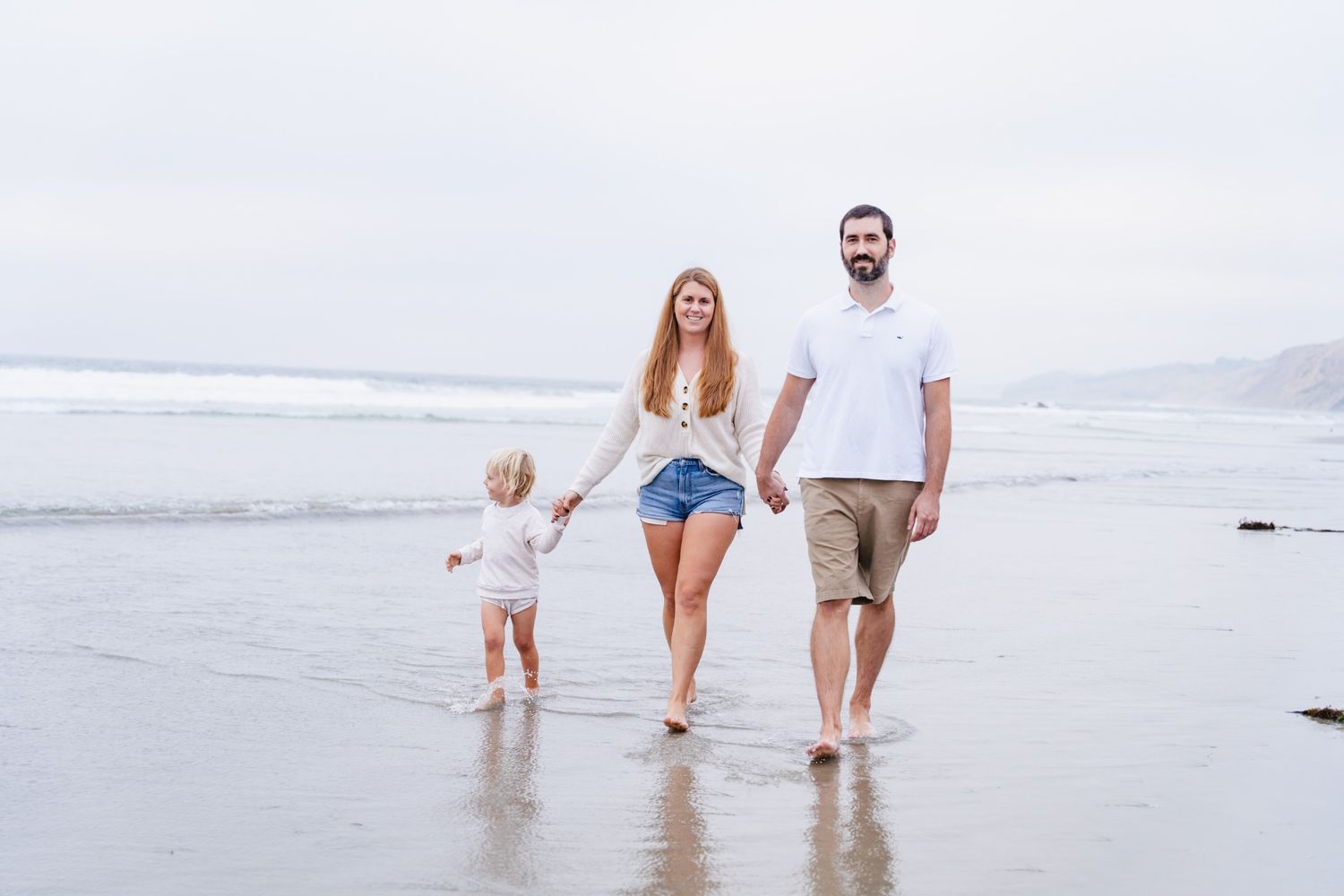 Young family walks barefoot on beach holding hands with toddler on cloudy day.