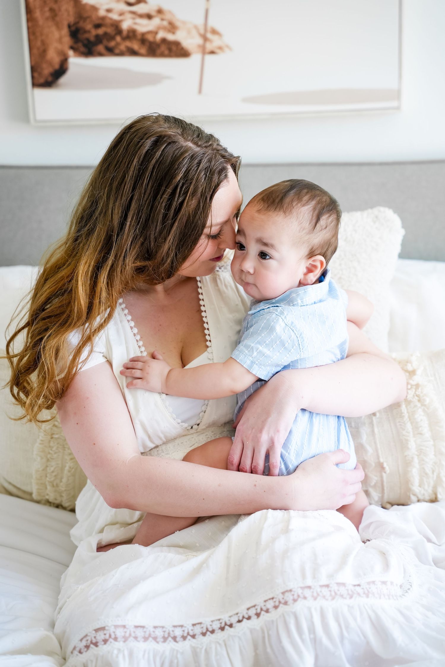 A mother in a white dress tenderly nuzzles her baby boy in a blue romper while sitting on a white bed.