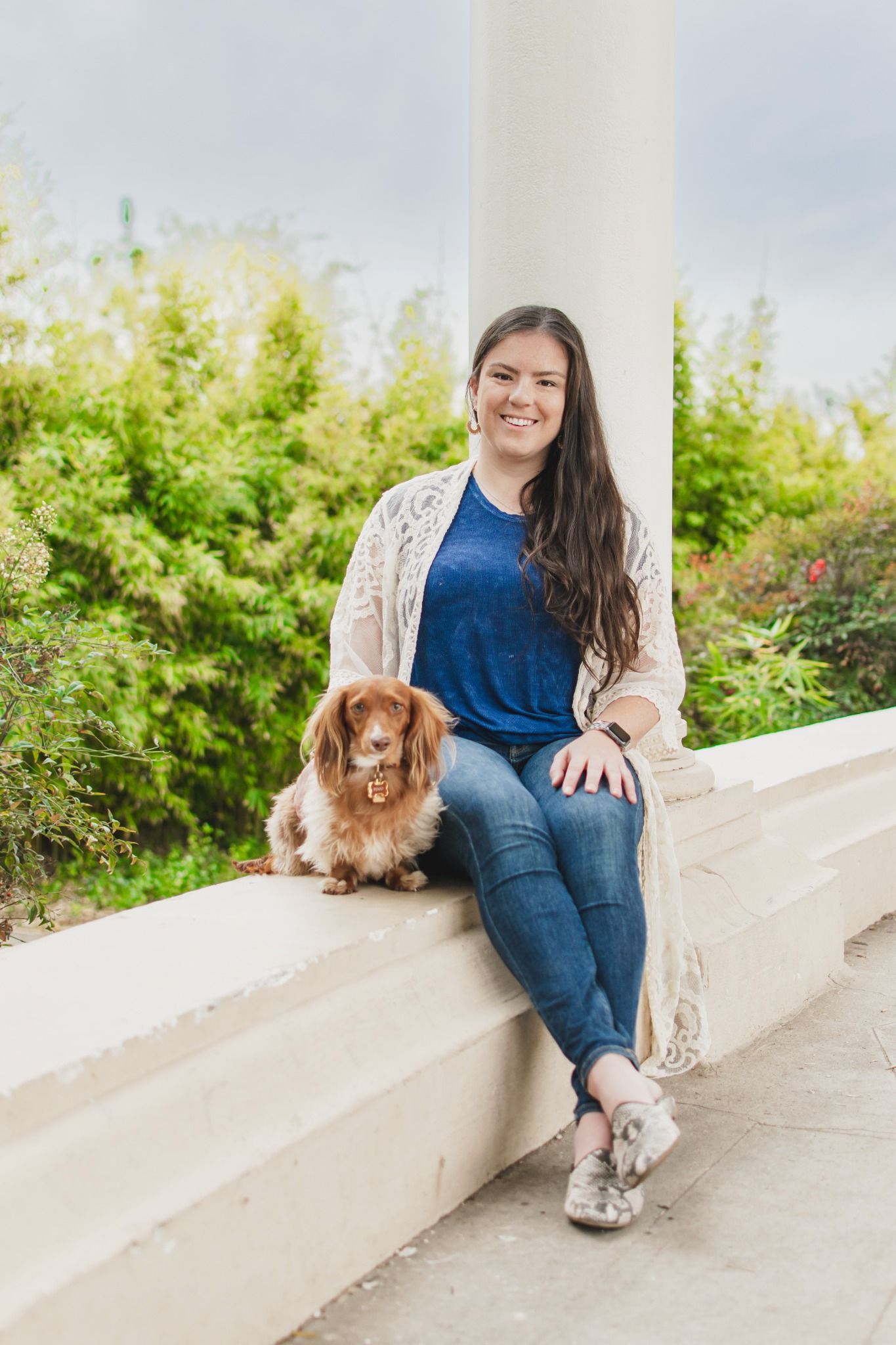 Young woman smiling with a cute Cavalier King Charles Spaniel dog, sitting outdoors on a white ledge with greenery.