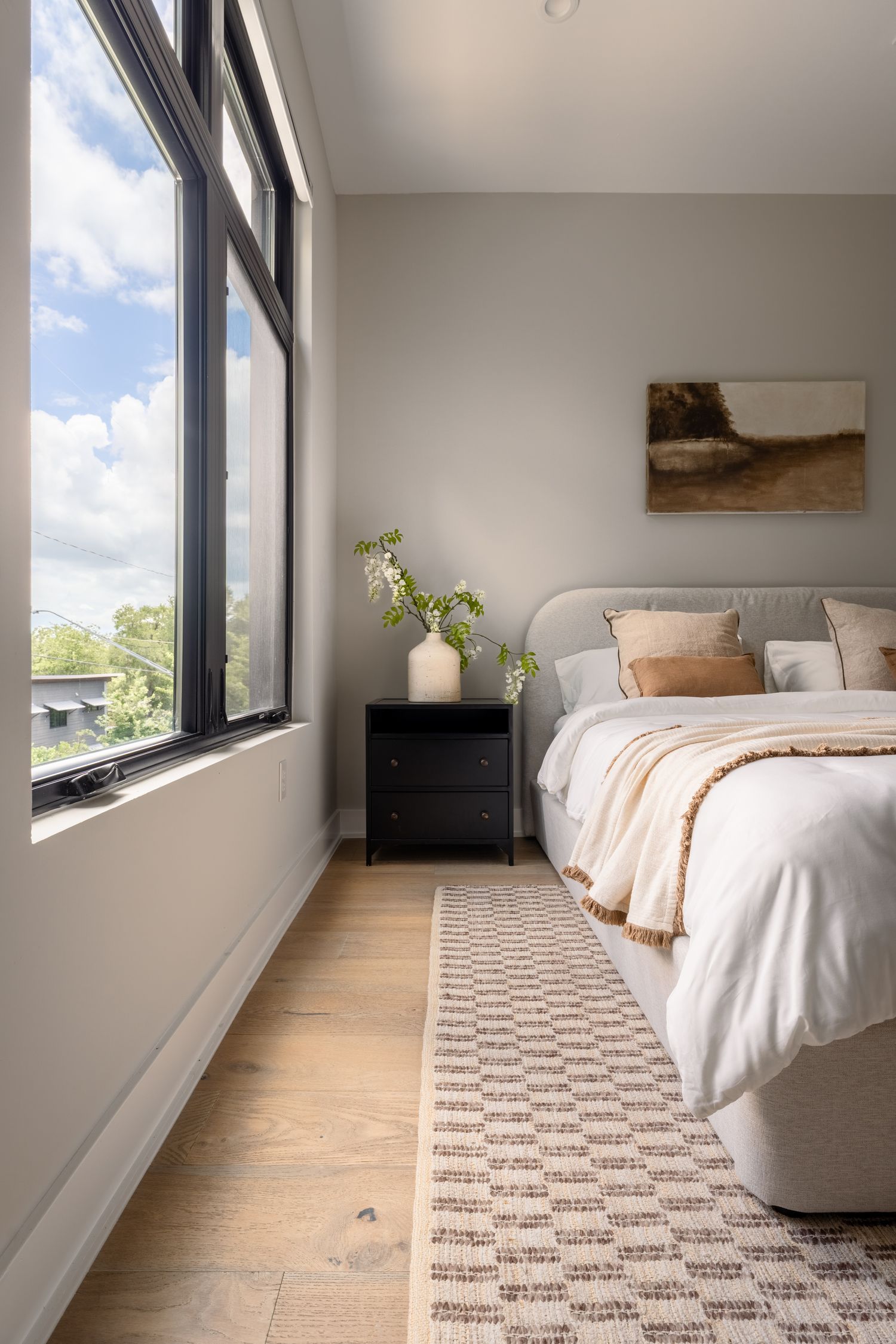 Modern bedroom with gray upholstered bed, black nightstand, patterned rug, and large windows with natural light.