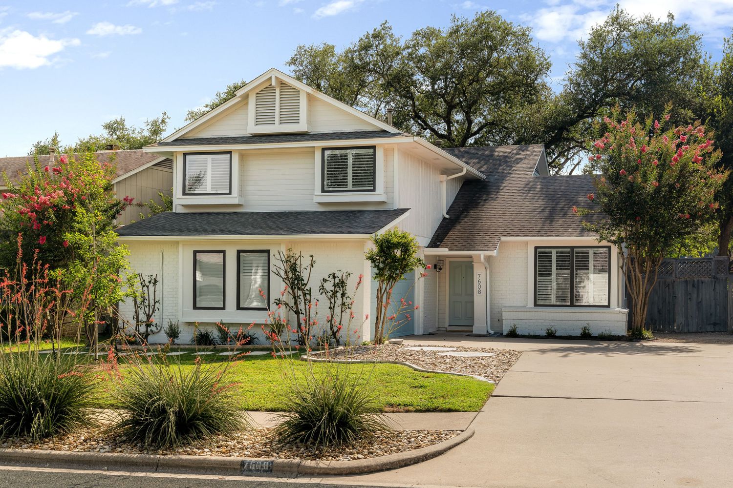 White two-story craftsman home with gray roof, manicured landscaping, and a wide driveway surrounded by mature oak trees.