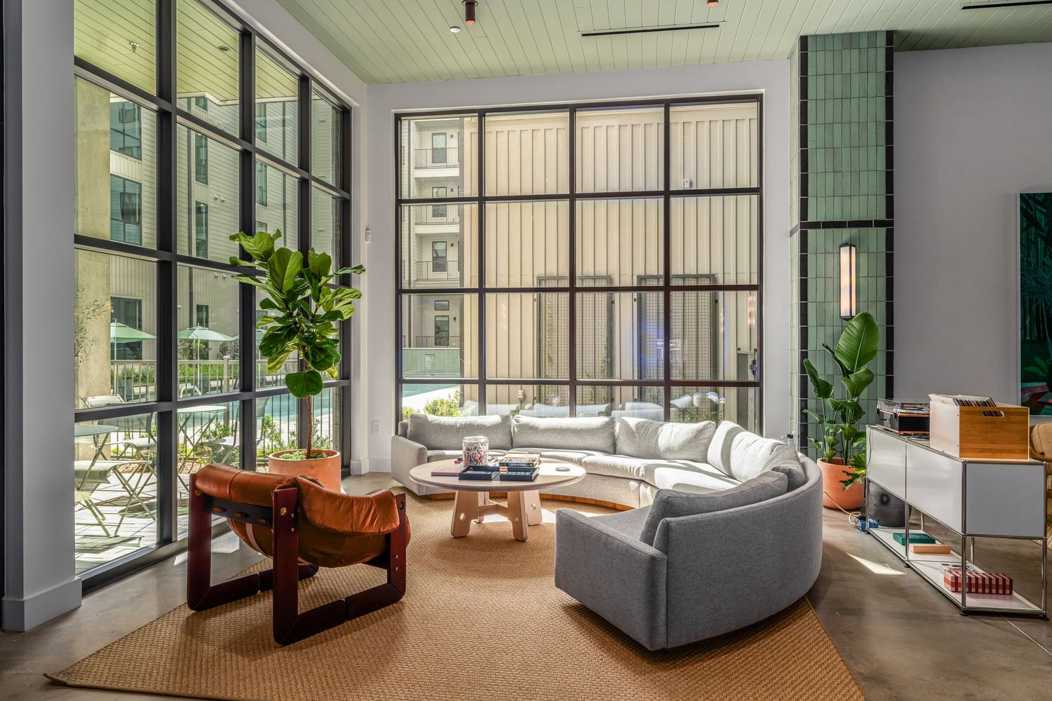 Modern living room with floor-to-ceiling windows, curved gray sofa, wood coffee table, and lush indoor plants in a bright space.