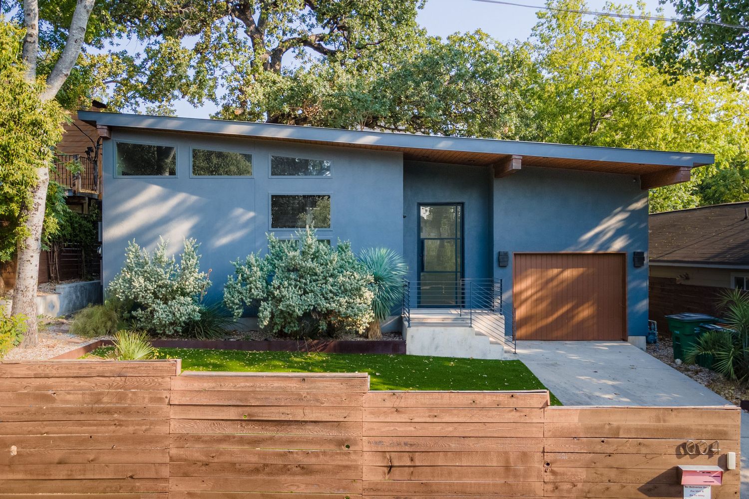 Modern mid-century blue home with wood garage door, cedar fence, and lush landscaping on a sunny day.