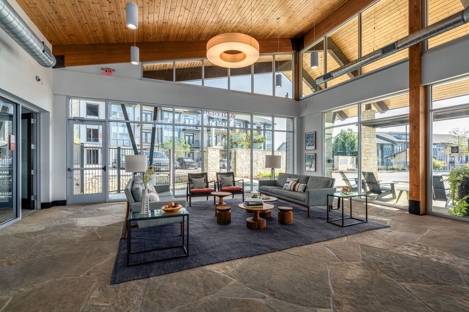 Modern apartment clubhouse lounge with high wood ceilings, large windows, stylish seating, and an open view of the outdoor courtyard.