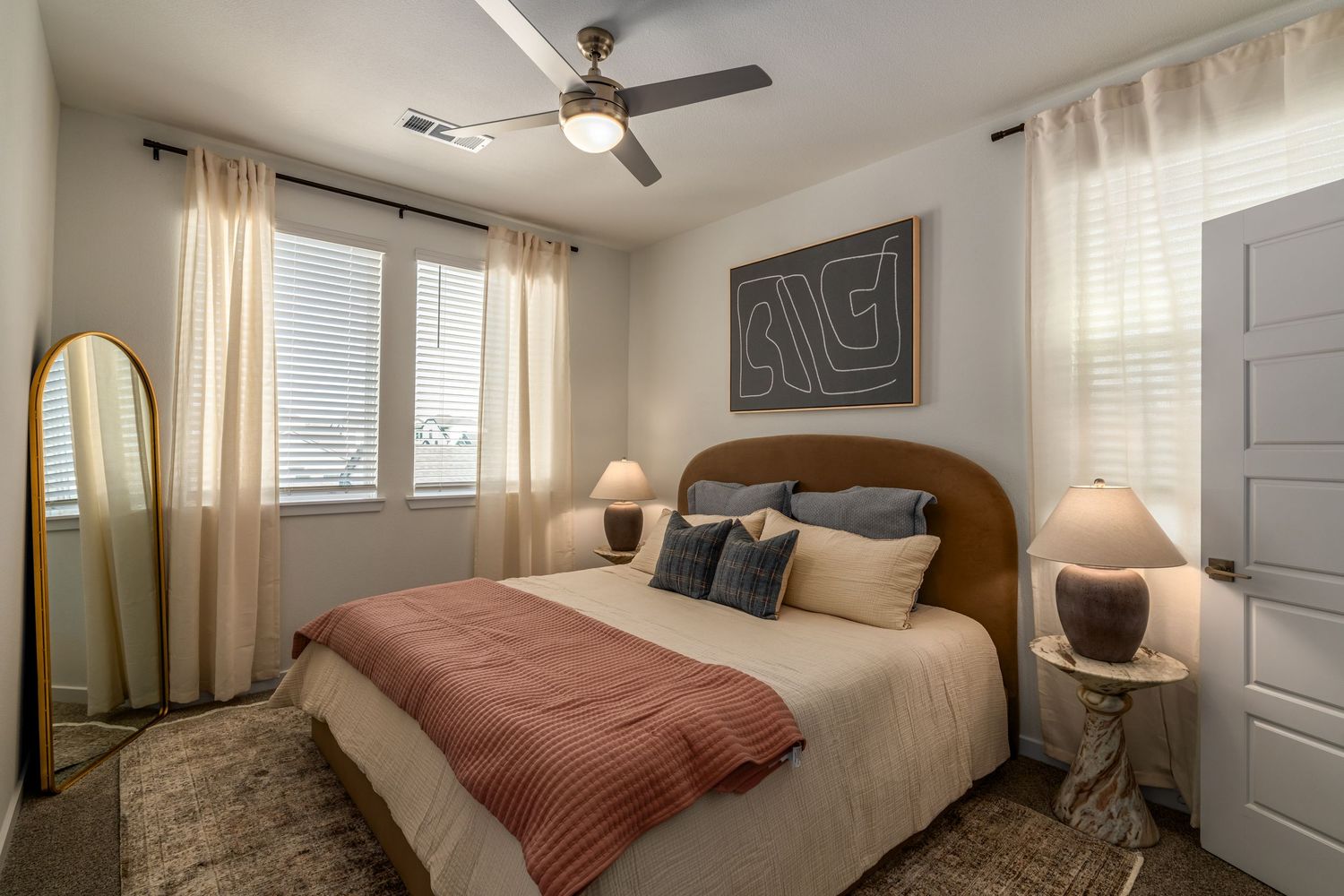 Cozy bedroom with queen bed, ceiling fan, warm lighting, decorative art, mirror, and sheer curtains in neutral tones.