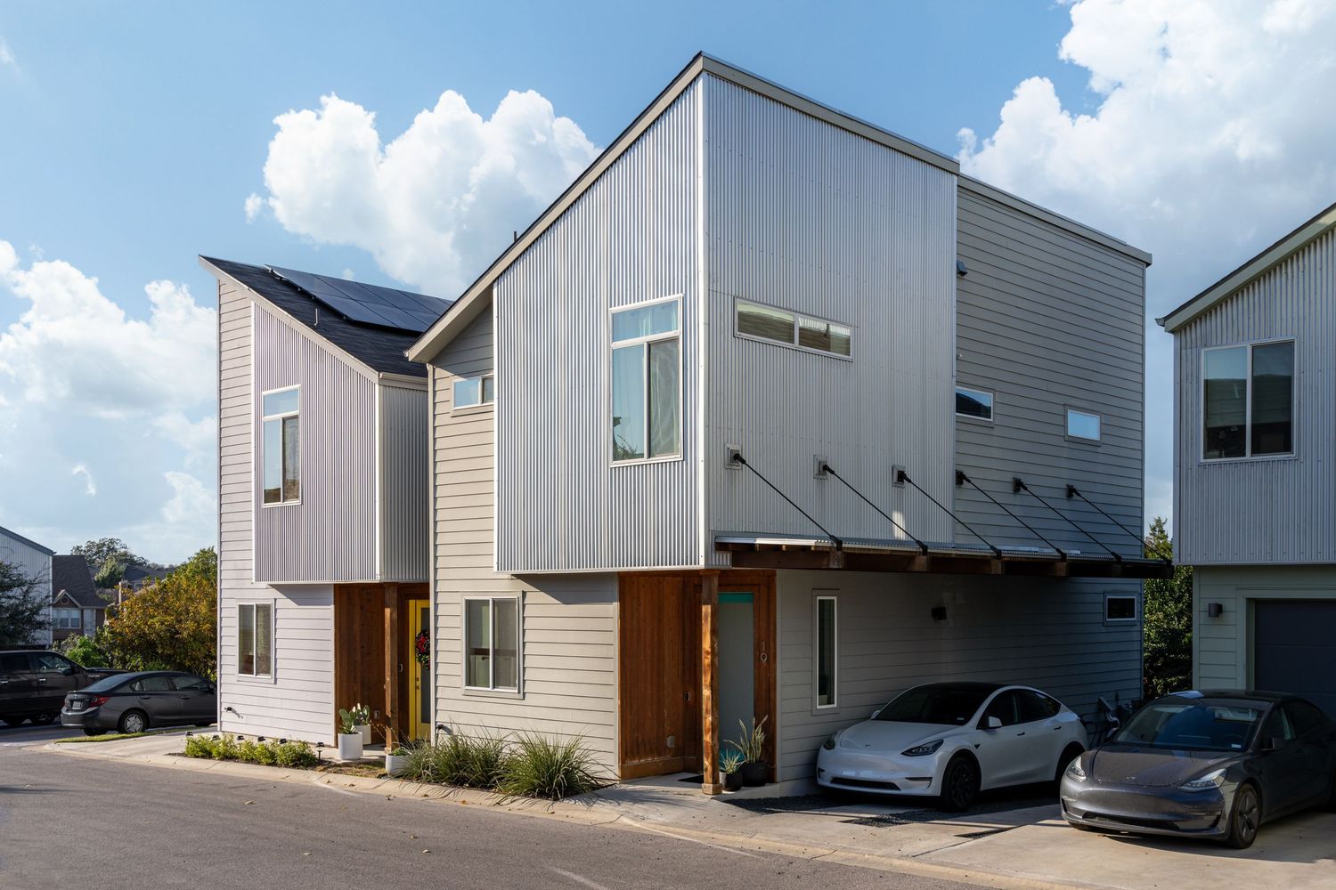 Modern two-story home with solar panels, mixed siding, wood accents, and a carport with Tesla parked outside.