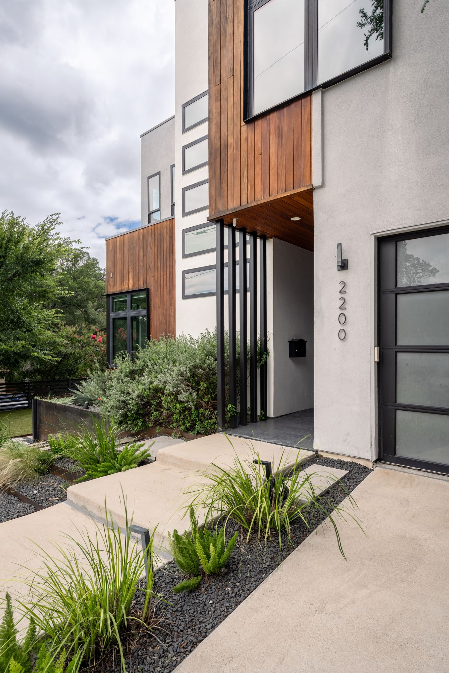 Modern two-story home exterior with wood accents, black steel entry, lush landscaping, and address number 2200 on stucco facade.