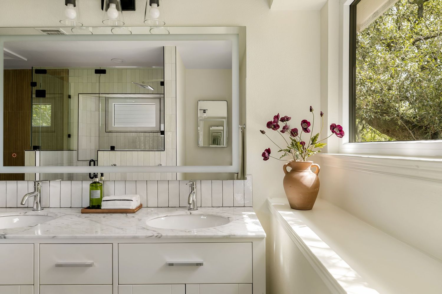 Bright bathroom with double white vanity, marble countertop, large mirror, and a vase of red flowers by a sunlit window.