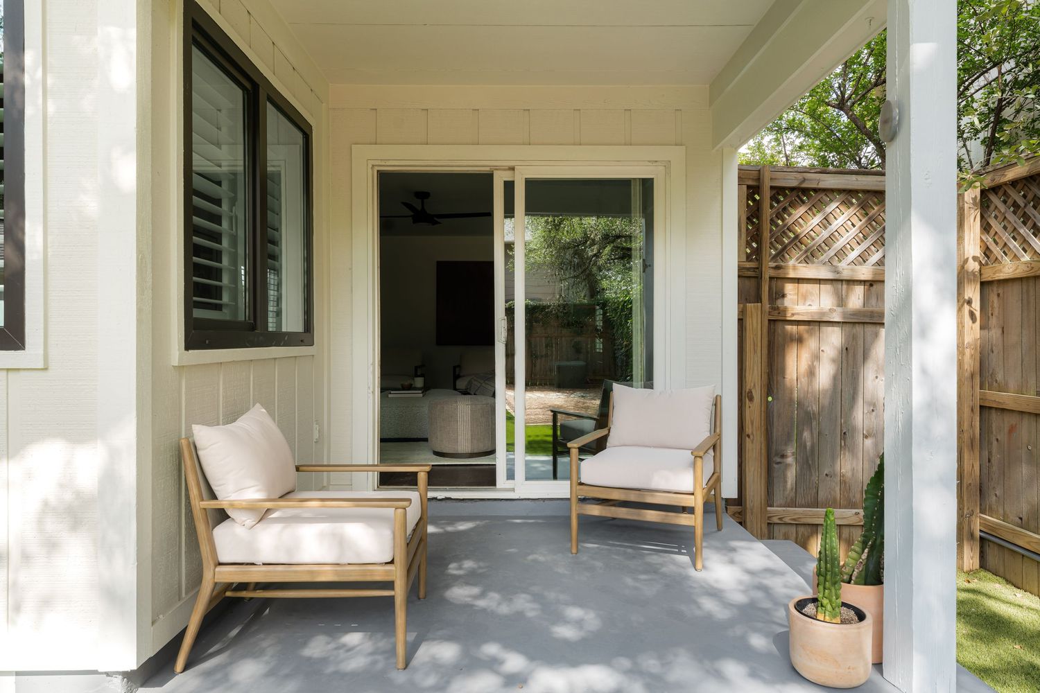 Cozy covered patio with two wooden chairs and white cushions beside a sliding glass door and wooden fence.