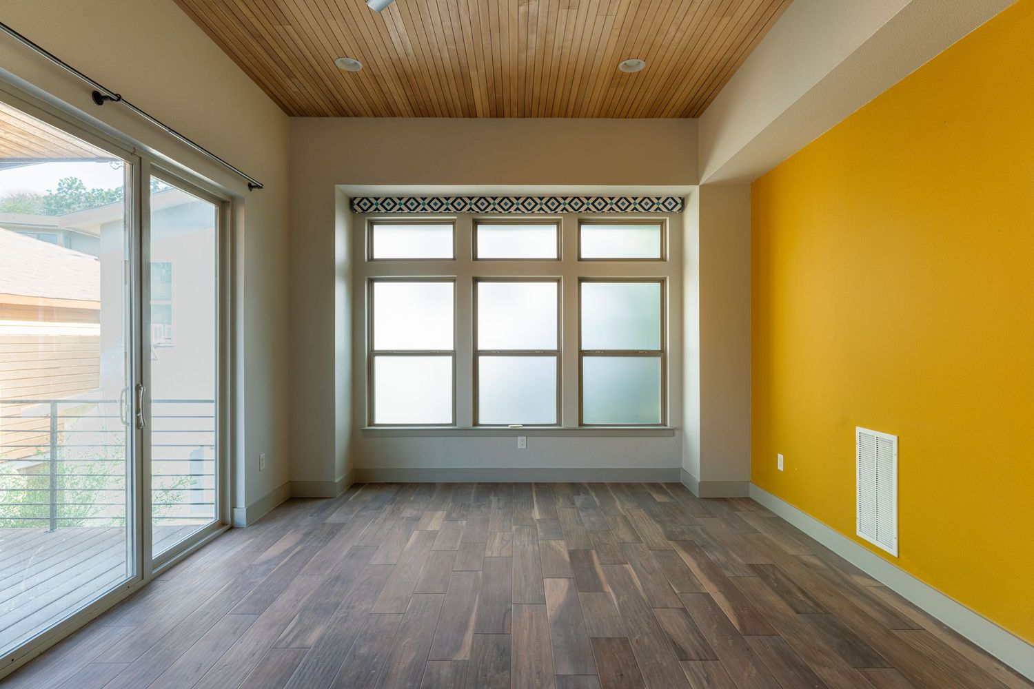 Empty room with yellow accent wall, wood ceiling, dark hardwood floors, and sliding glass door letting in natural light.