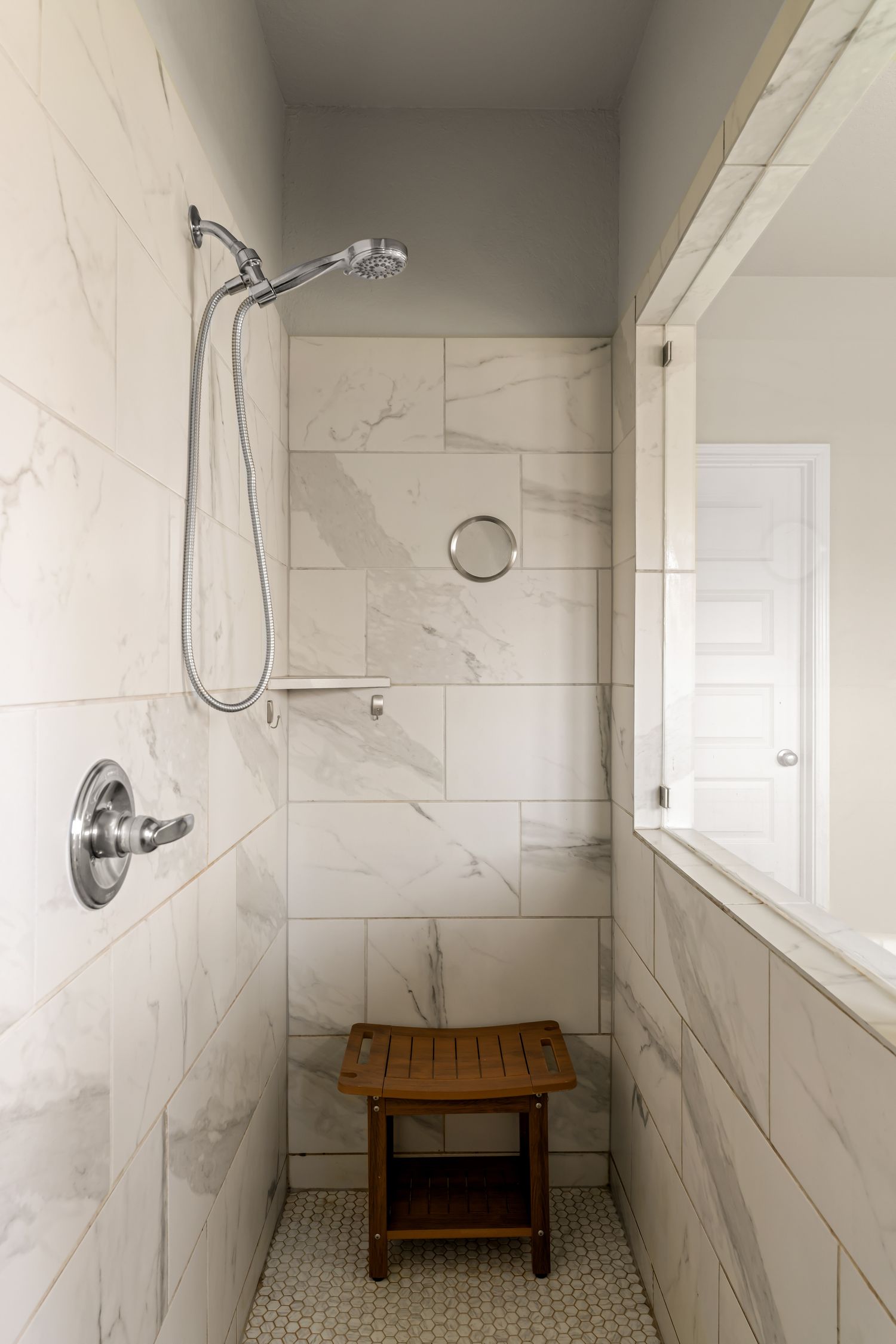 Walk-in shower with marble tile walls, chrome rainfall showerhead, handheld sprayer, and wooden teak bench.