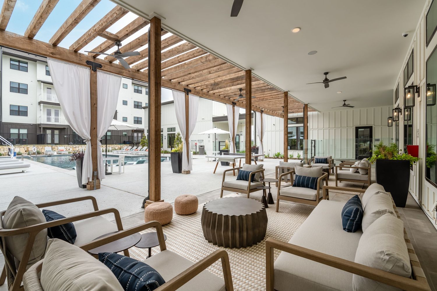 Elegant outdoor lounge area with wood pergola, white sofas, and ceiling fan overlooking a resort-style pool.