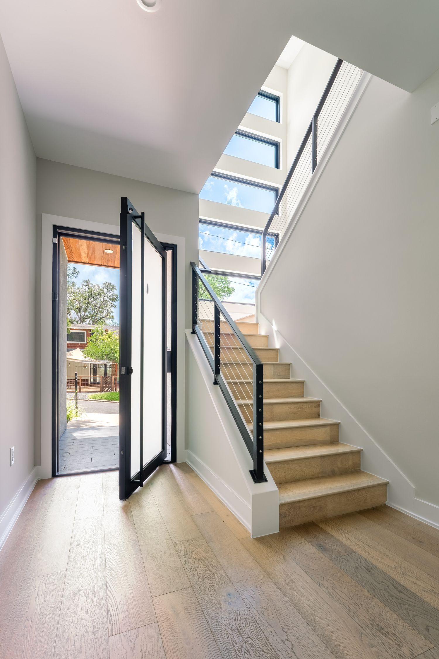 Modern home entryway with open front door, light wood staircase, black metal railing, and skylights flooding natural light.