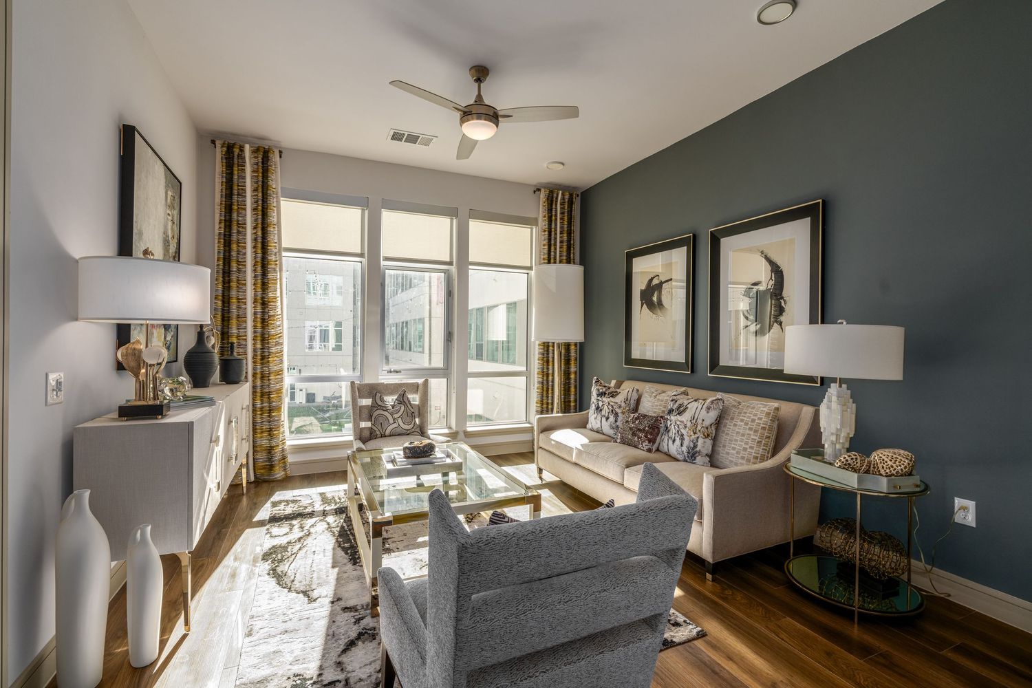 Modern living room with gray sofa, glass coffee table, hardwood floors, and large windows with gold curtains.