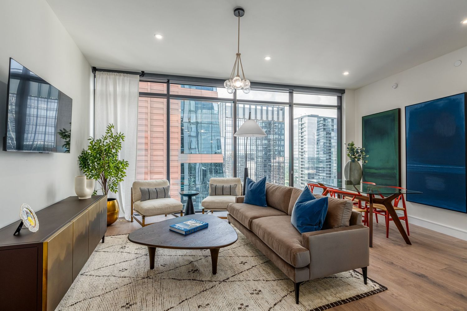 Modern living room with floor-to-ceiling windows, city views, gray sofa with blue pillows, and elegant chandelier lighting.