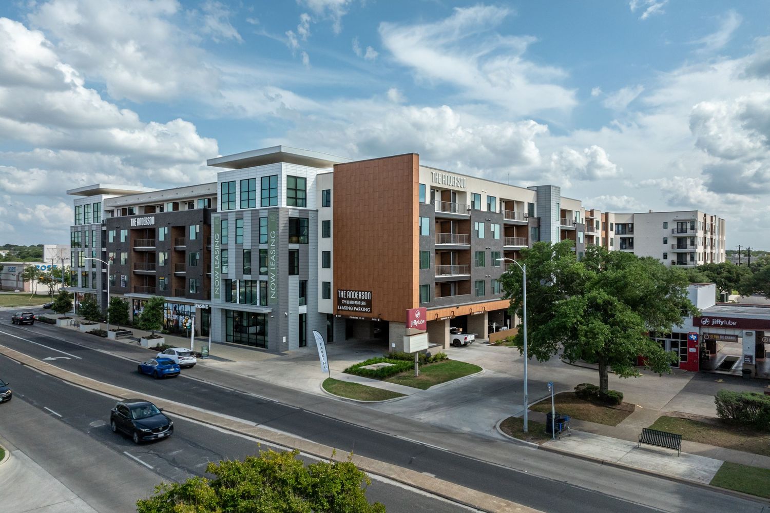 Modern mixed-use apartment building with ground-floor retail on a sunny urban street corner with tree-lined sidewalks.