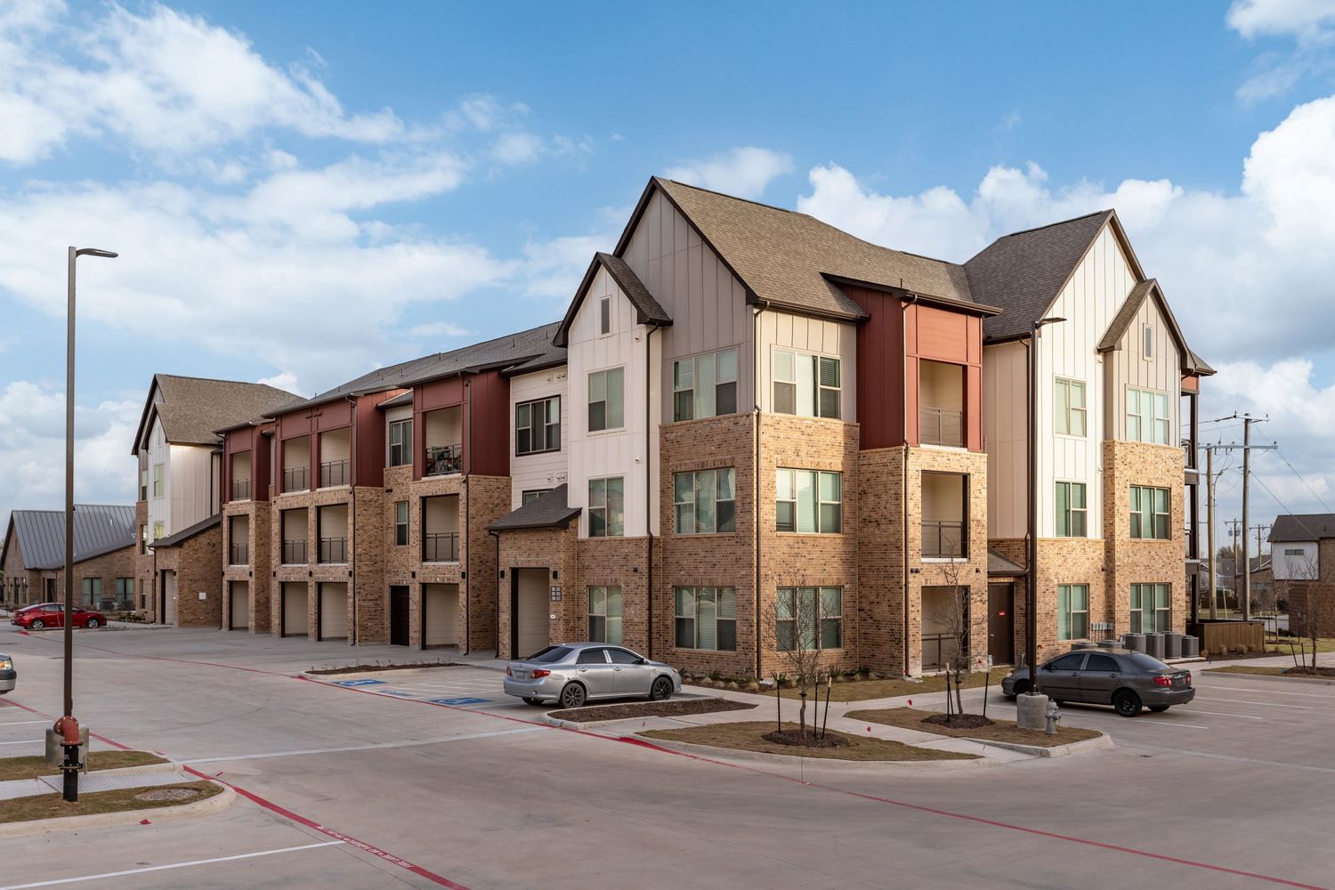 Modern three-story apartment complex with brick and stone exterior, balconies, and parking along a quiet suburban street.