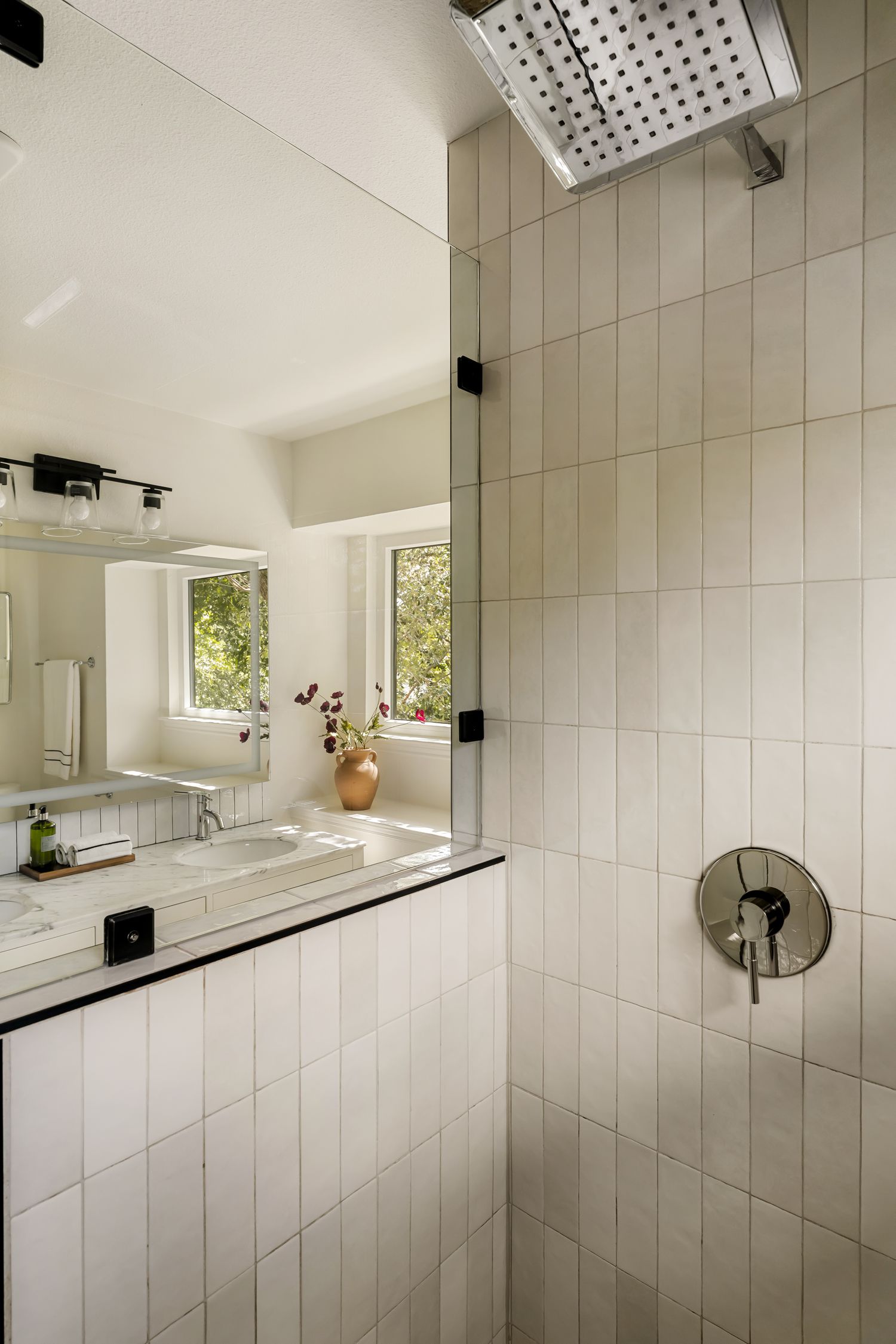 Modern bathroom with white subway tiles, rainfall shower head, marble vanity, and a window view of lush greenery.