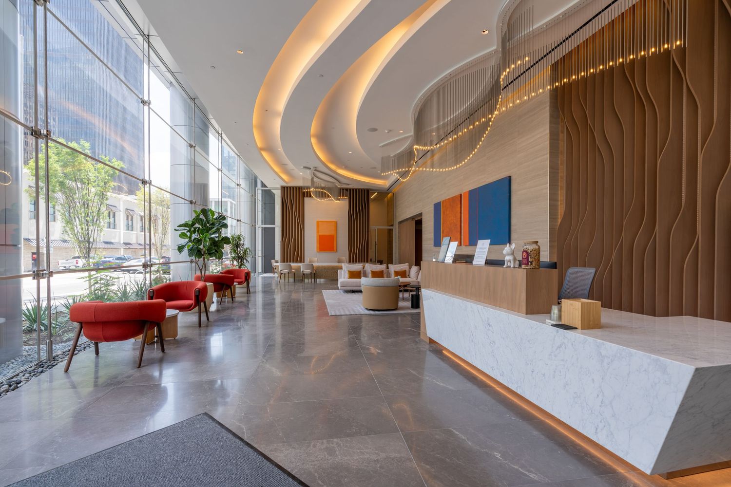 Modern hotel lobby with curved ceiling lights, marble reception desk, red accent chairs, and warm wood wall panels.