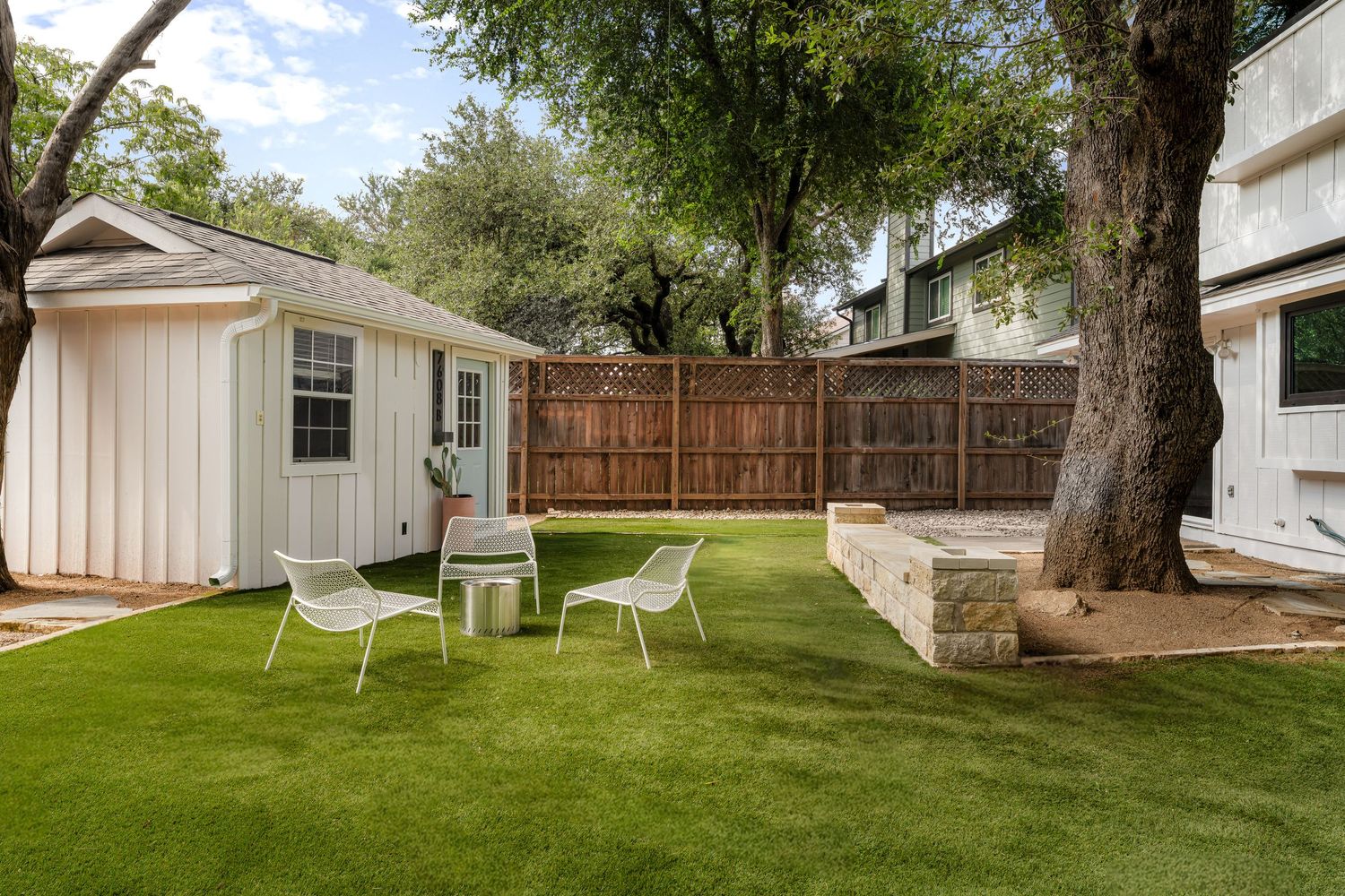 Backyard with lush green artificial turf, white patio chairs, a storage shed, wooden privacy fence, and mature shade trees.