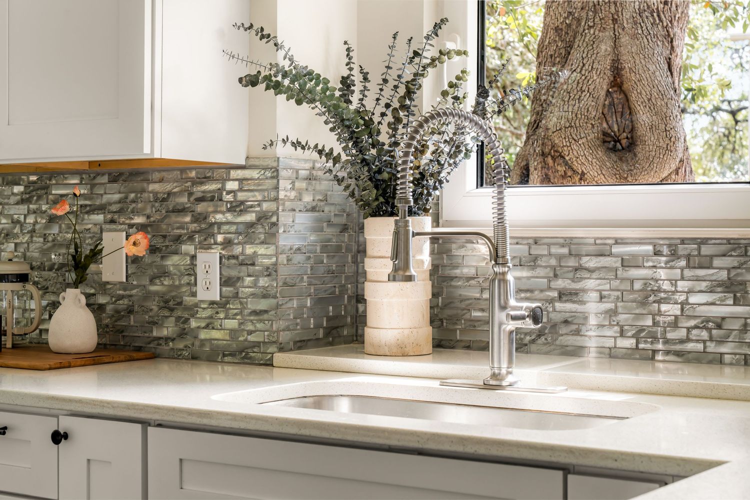 Modern kitchen sink with chrome faucet, gray mosaic tile backsplash, white quartz countertop, and decorative plants by a window.