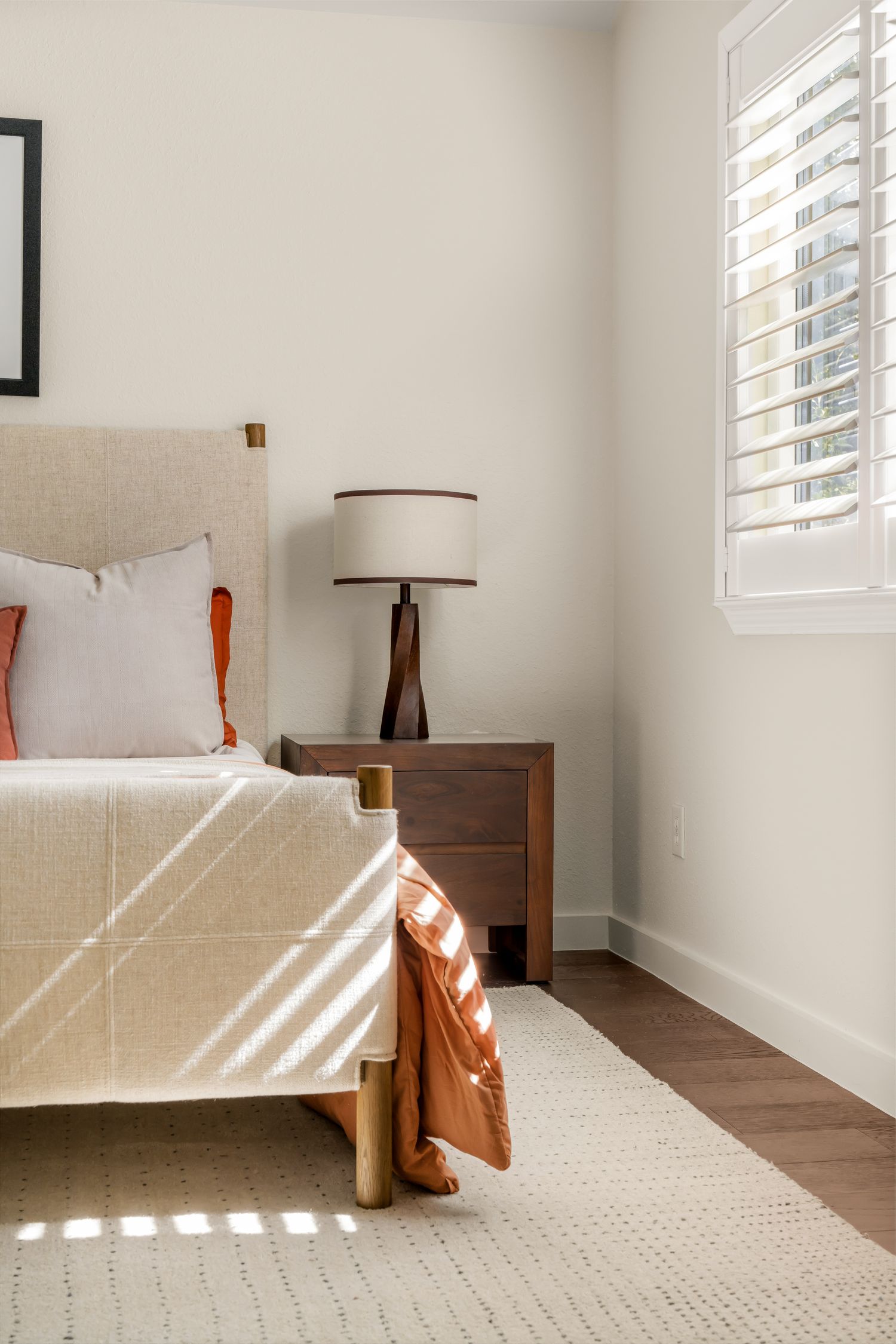 Bright bedroom with beige upholstered bed, terracotta pillows, wood nightstand, lamp, and plantation shutters on hardwood floors.