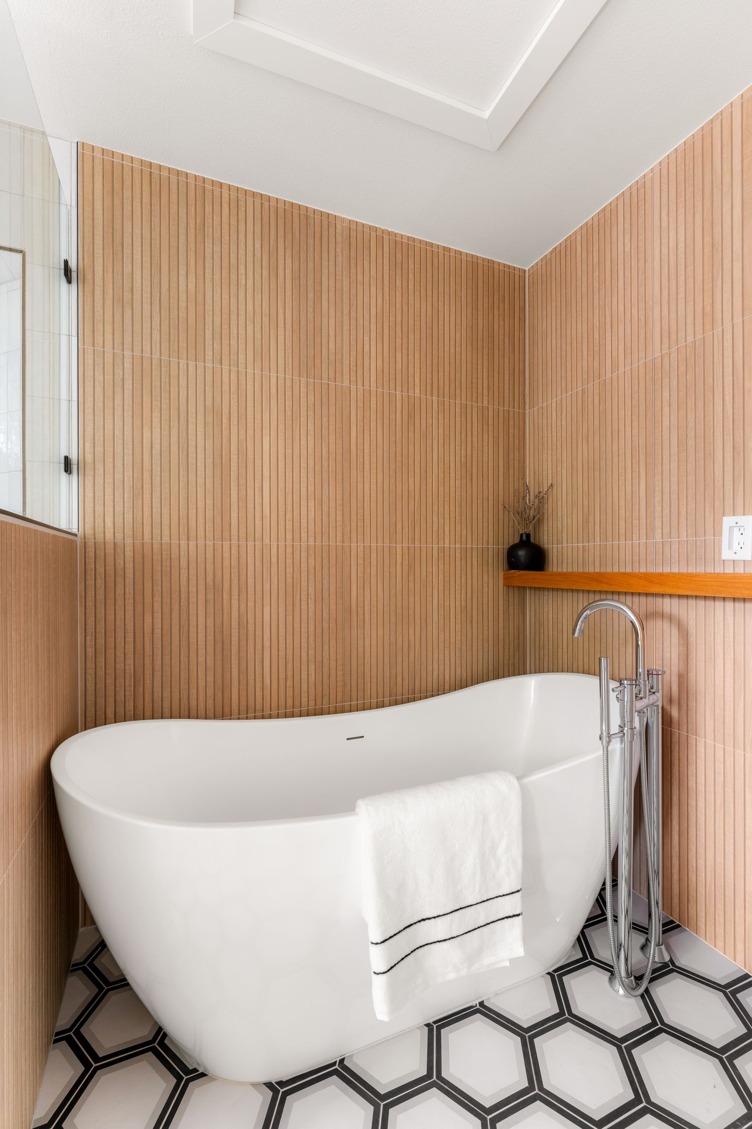 Freestanding white bathtub on hexagon tile floor with warm terracotta fluted wall tiles and wooden shelf in modern bathroom.