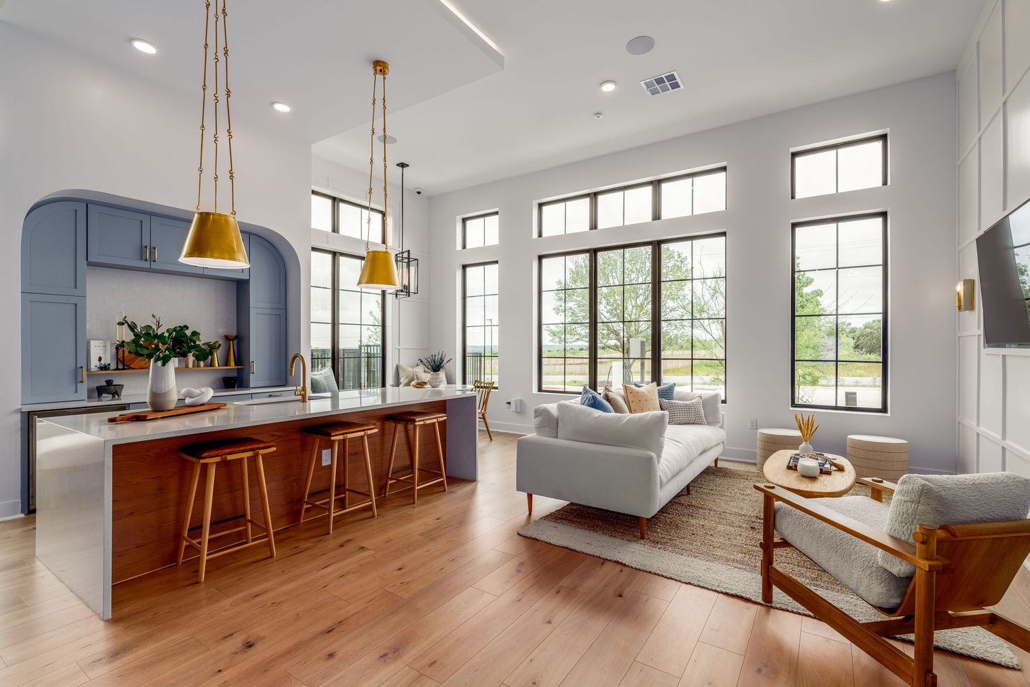 Open-concept living room and kitchen with hardwood floors, blue arched cabinetry, gold pendant lights, and large windows.