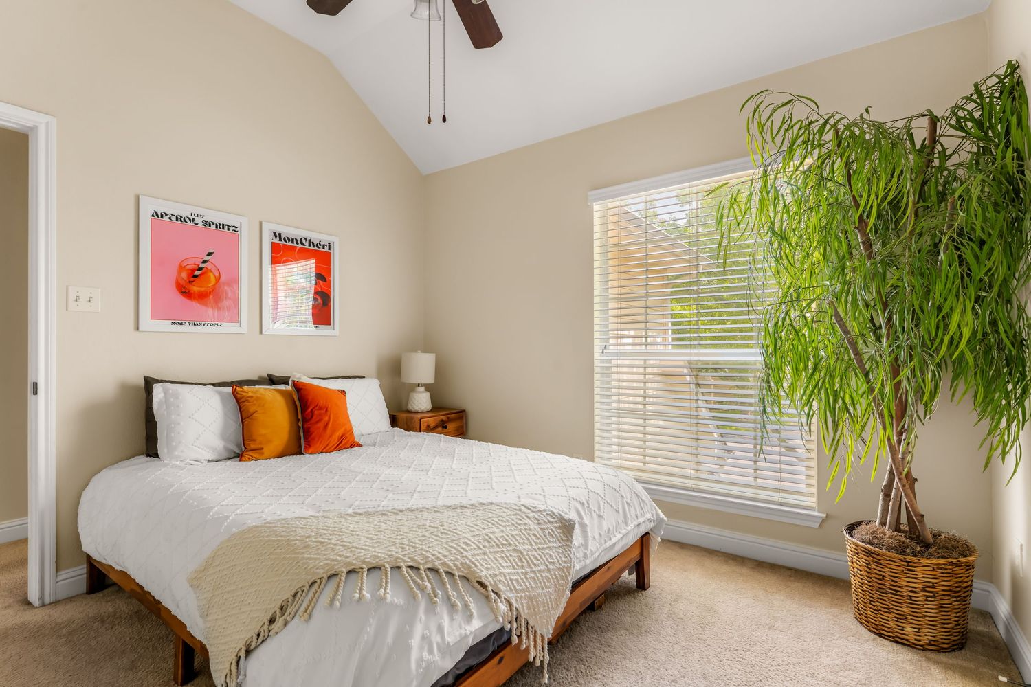 Cozy bedroom with vaulted ceiling, ceiling fan, queen bed with orange pillows, large window, and tall potted plant.