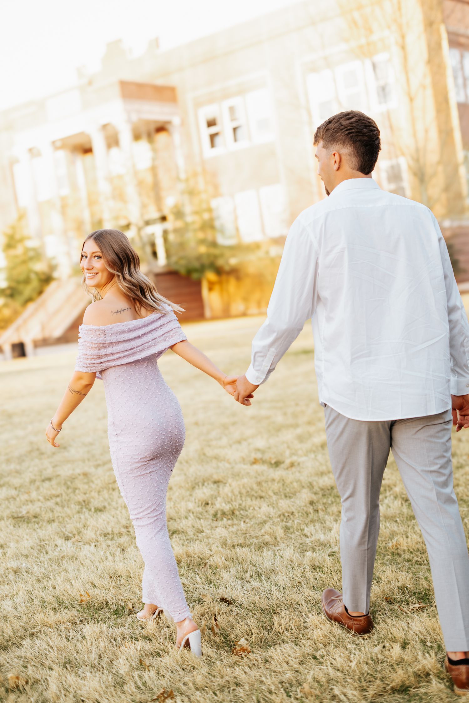 Couple holding hands walking across green lawn near elegant building in warm sunset light.