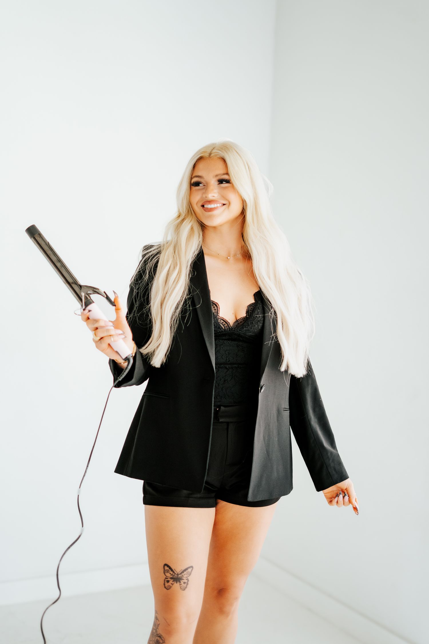 Smiling blonde woman in black blazer holding a curling iron with butterfly leg tattoo.