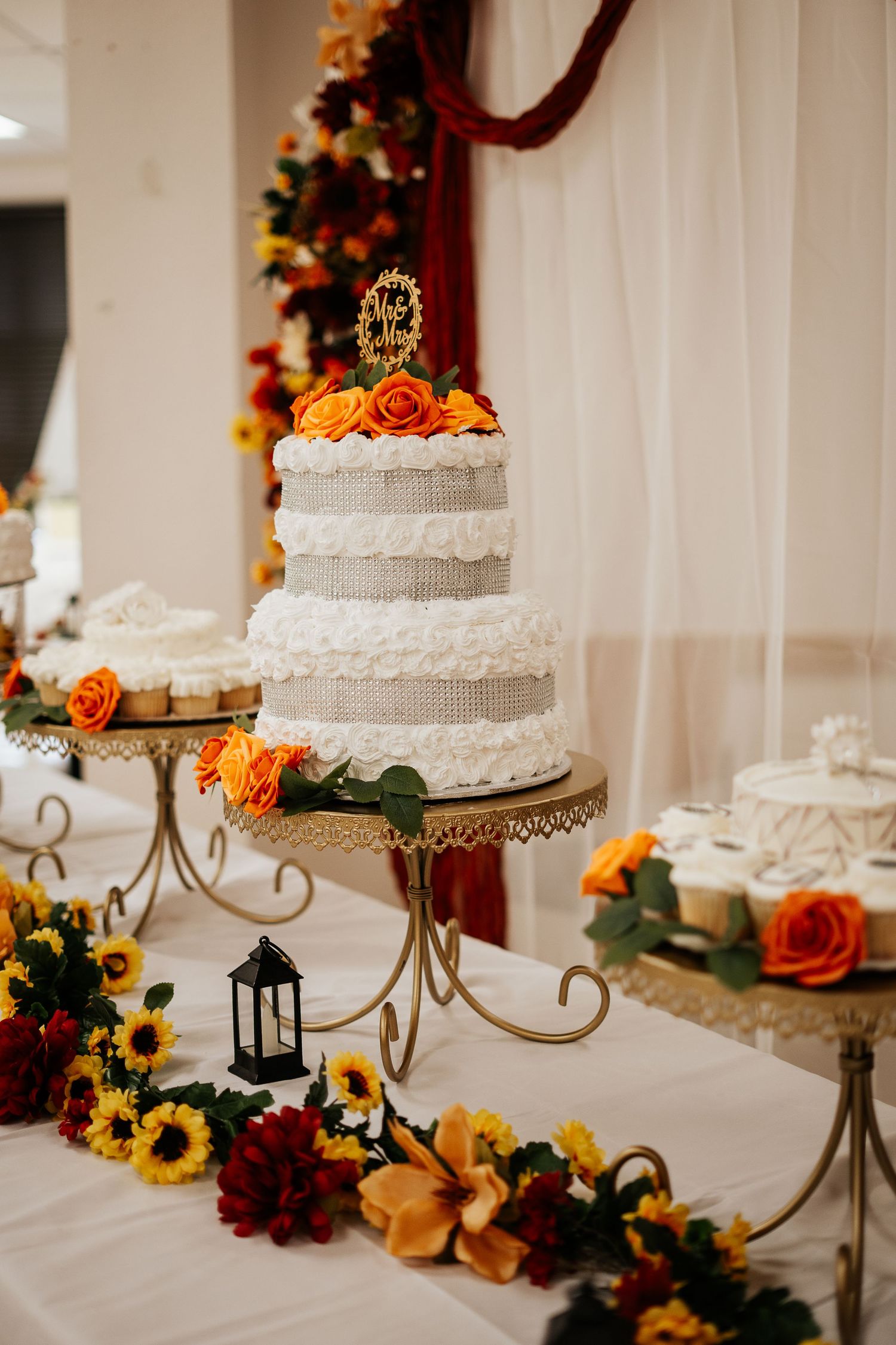 Three-tier white wedding cake with orange roses and Mr and Mrs topper surrounded by sunflowers and fall florals.