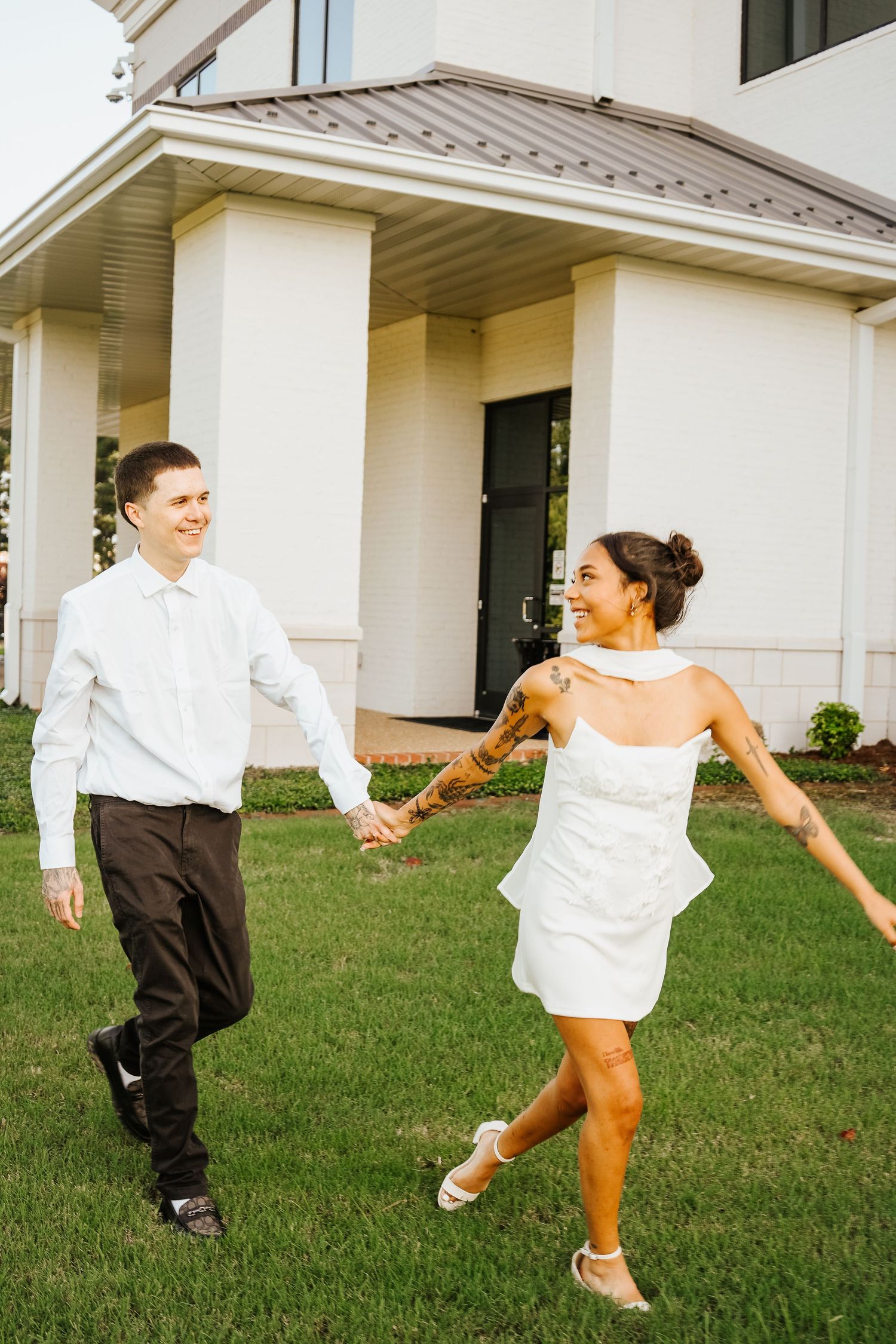 Happy couple holding hands and dancing on green grass outside a white building during their wedding day.