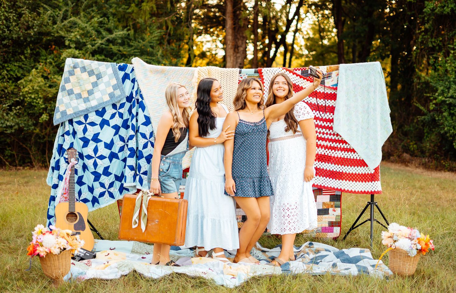 Four women in summer dresses posing with colorful quilts hanging outdoors in a grassy field.