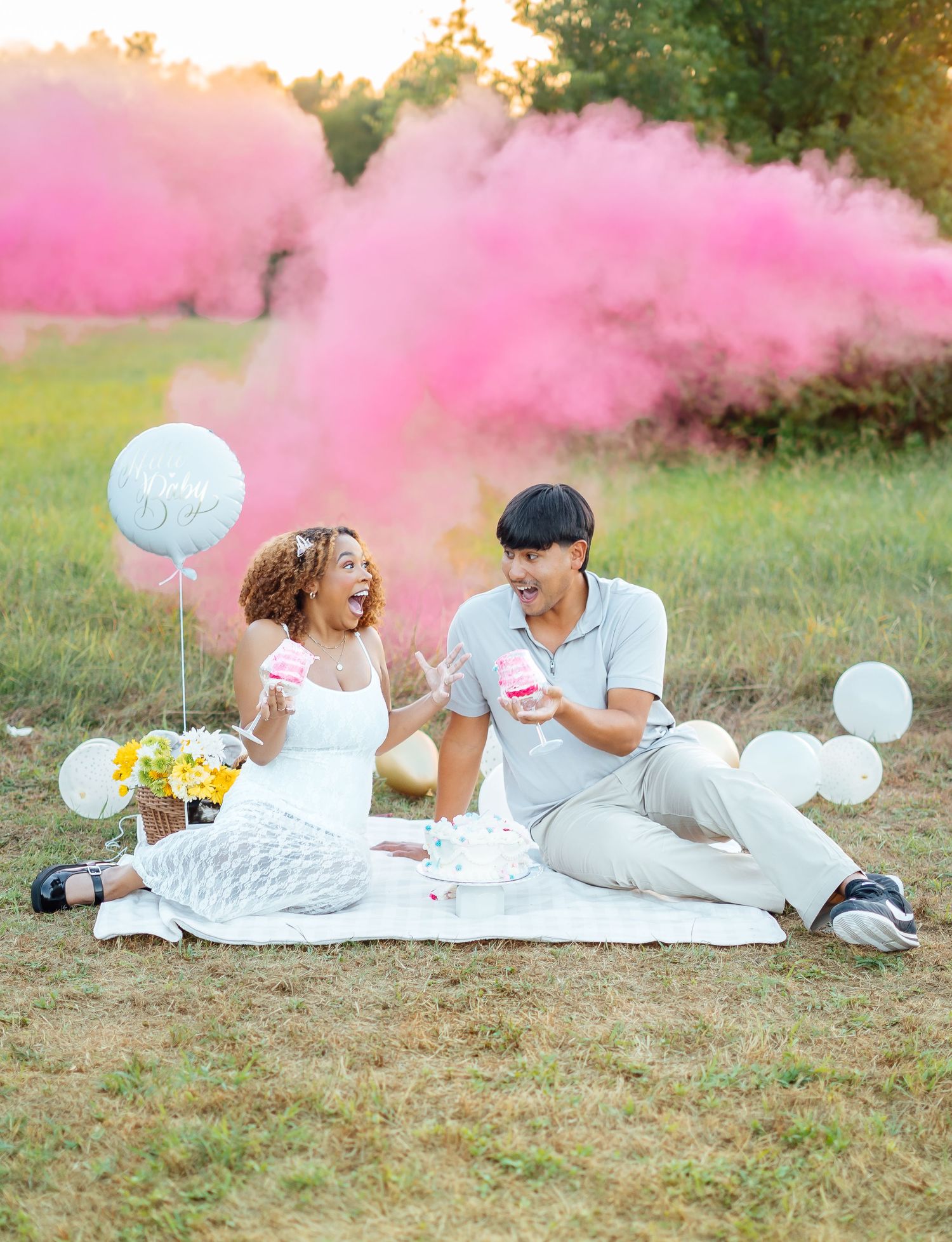 Couple sitting on blanket outdoors with pink gender reveal smoke bomb and white balloons.