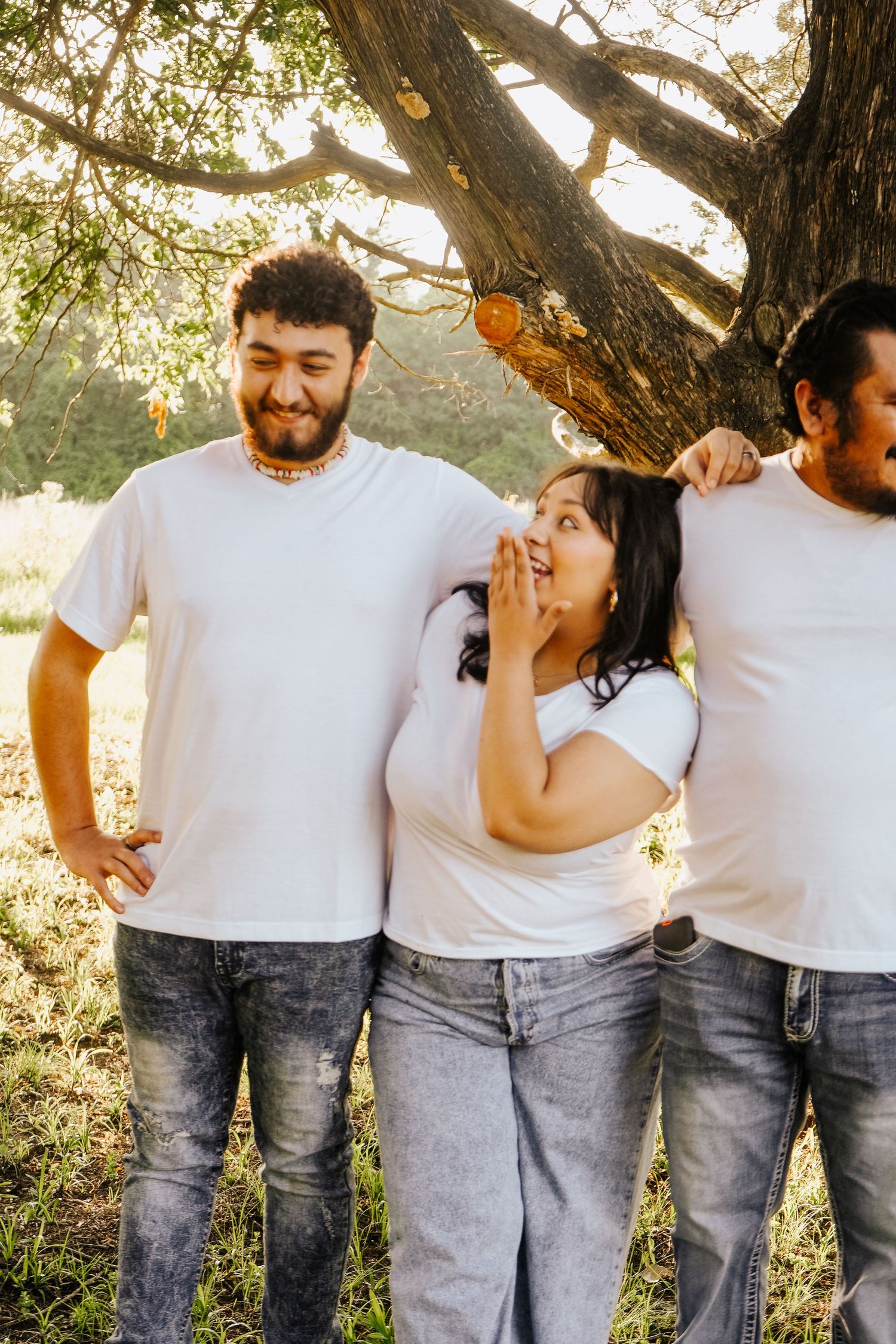 Three siblings in white tees sharing a candid laughing moment under a tree outdoors.